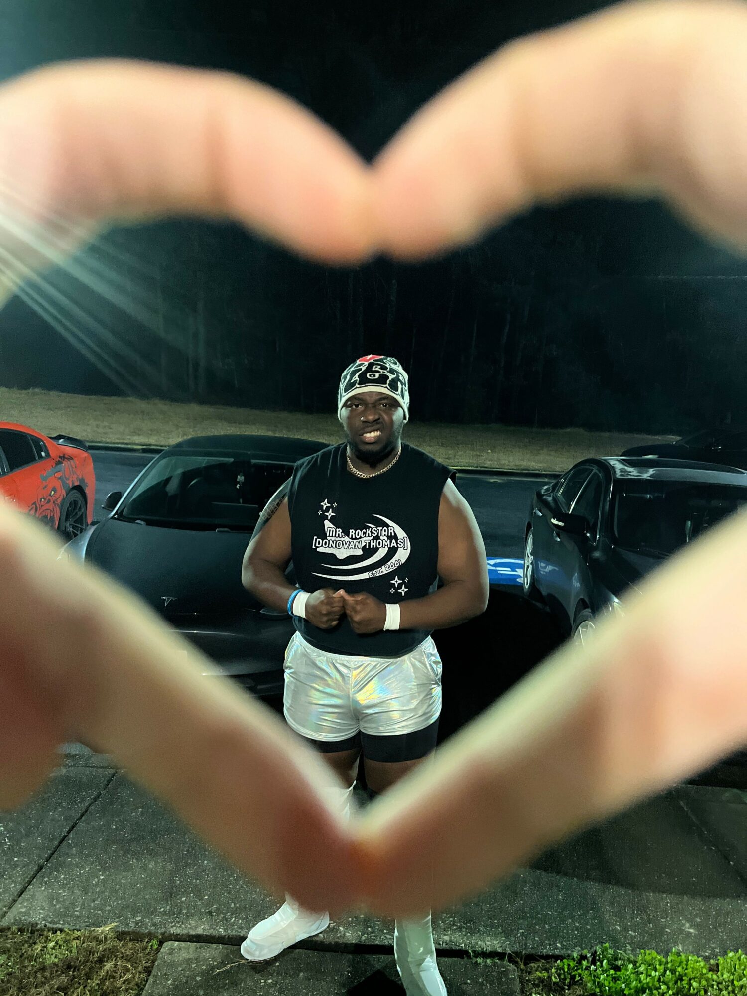 Person standing outdoors at night, wearing a cap, sleeveless shirt, and shorts, surrounded by parked cars.