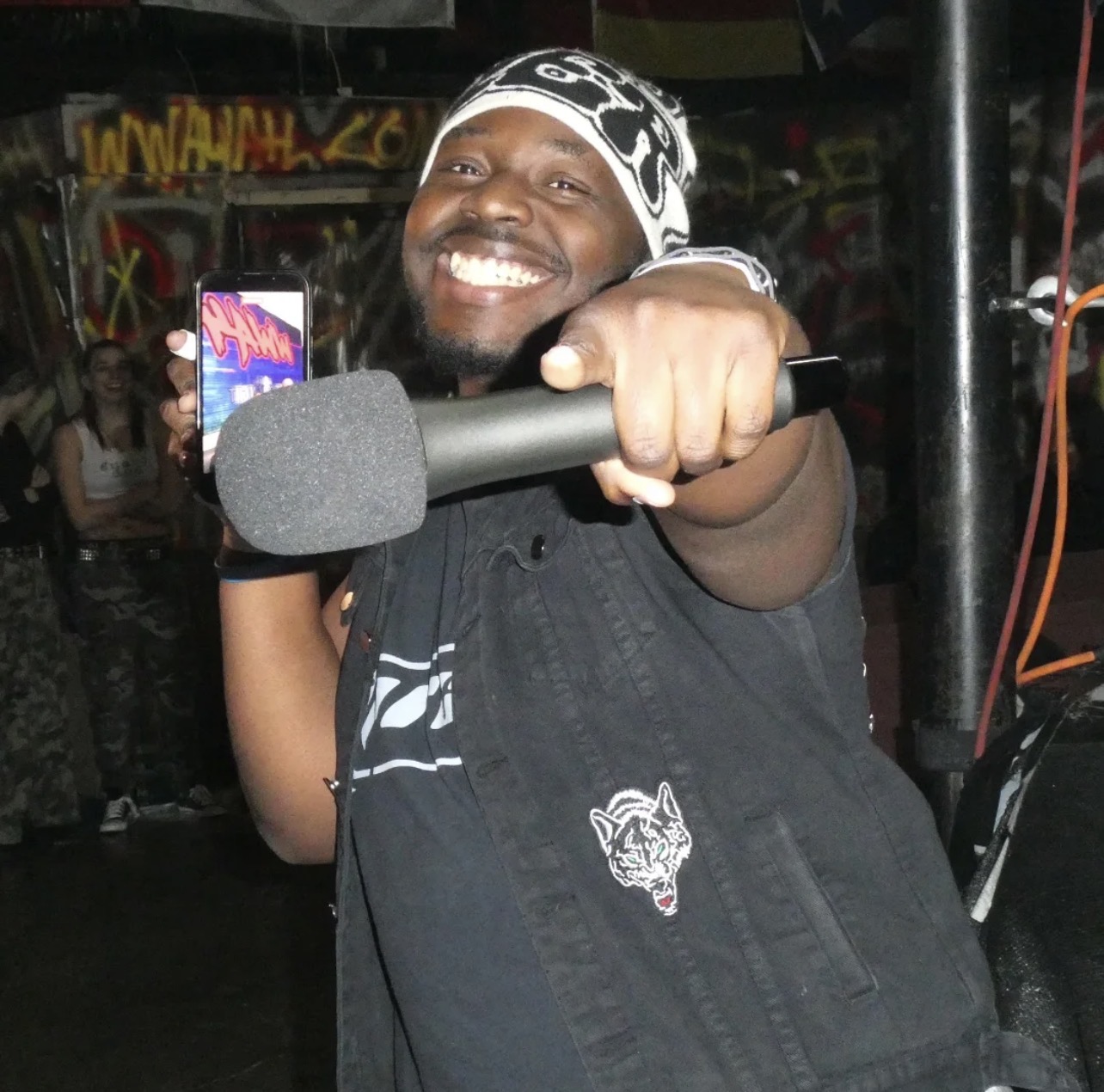 Person smiling and pointing microphone towards camera in a lively environment.