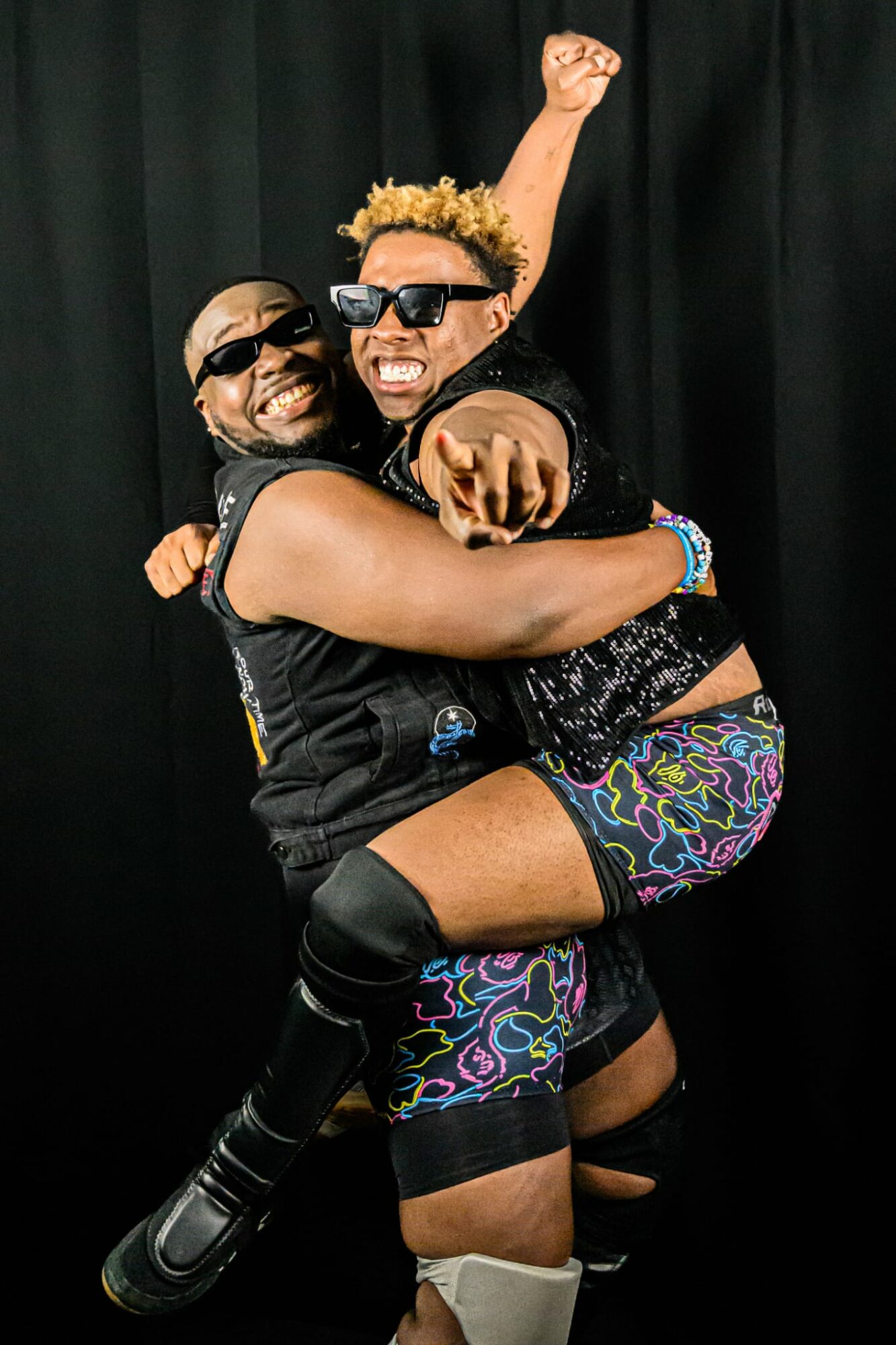 Two people smiling, one lifting the other, both wearing sunglasses and colorful clothing, black background.