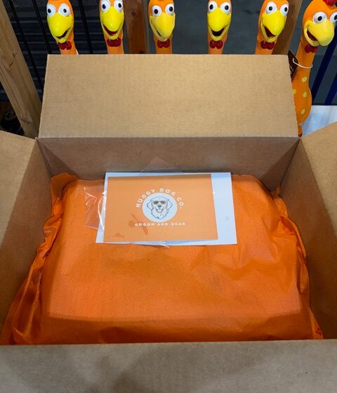 Open cardboard box with orange wrapped item inside, label with a bear logo on top, colorful giraffe toys in background.