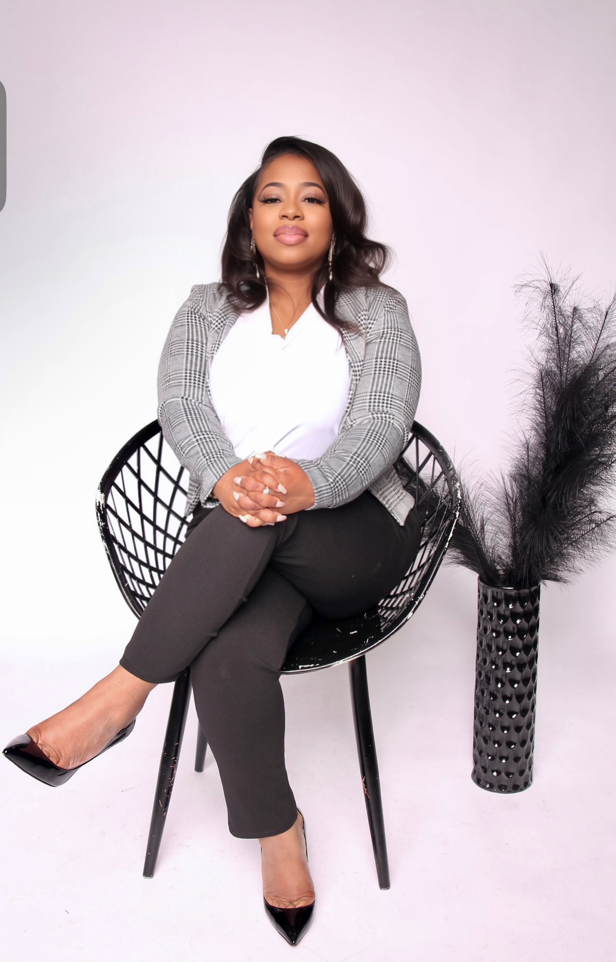 Woman sitting on a black chair with crossed legs, smiling, wearing a gray blazer, white blouse, black pants, and heels.