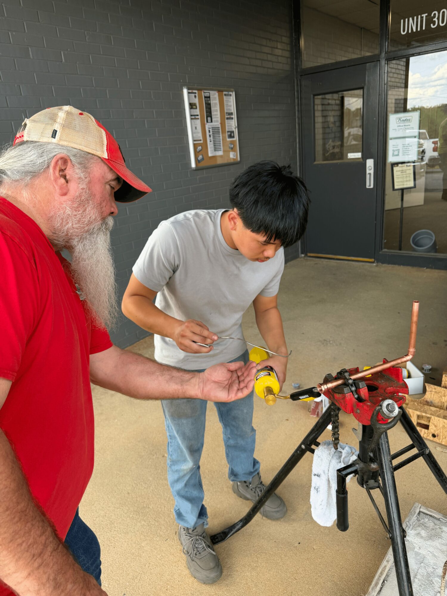 Two people working on a red and black device outdoors near a building entrance, with a bulletin board and glass door in background.