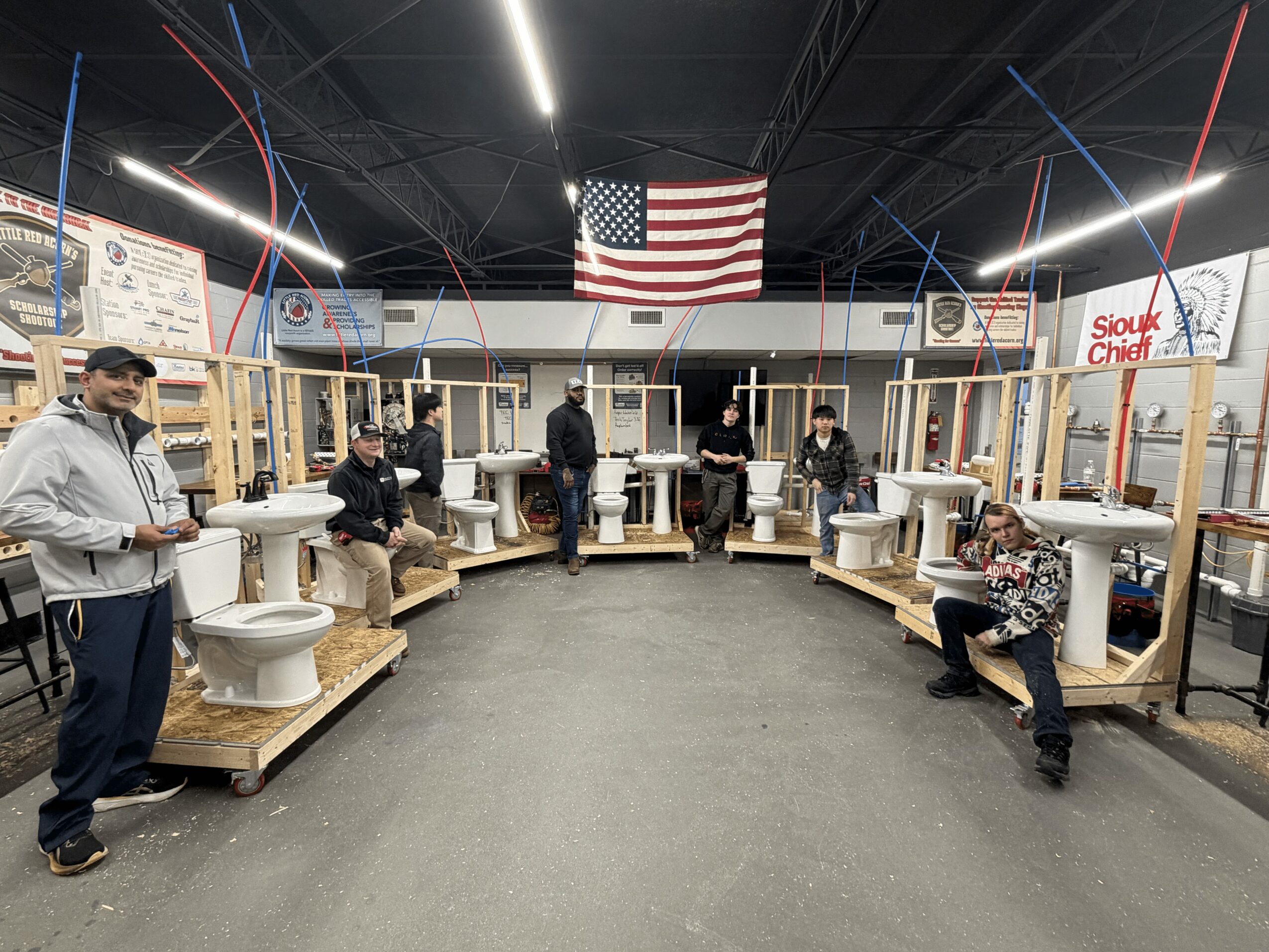 Group of people in a workshop with toilets and wooden structures, American flag hanging above, industrial ceiling with lights.