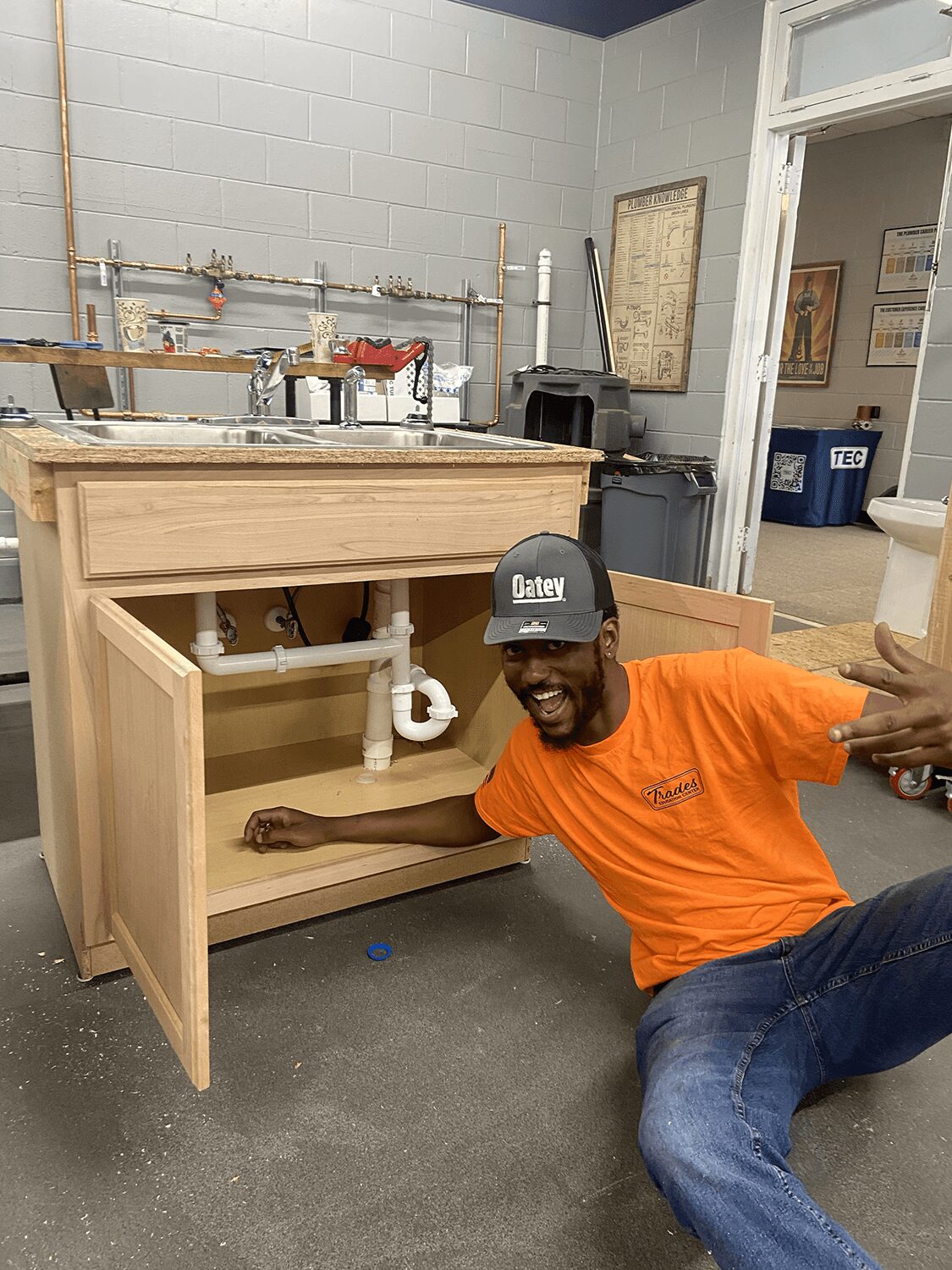 Man in orange shirt and cap sitting on the floor next to a wooden cabinet under a sink, smiling.