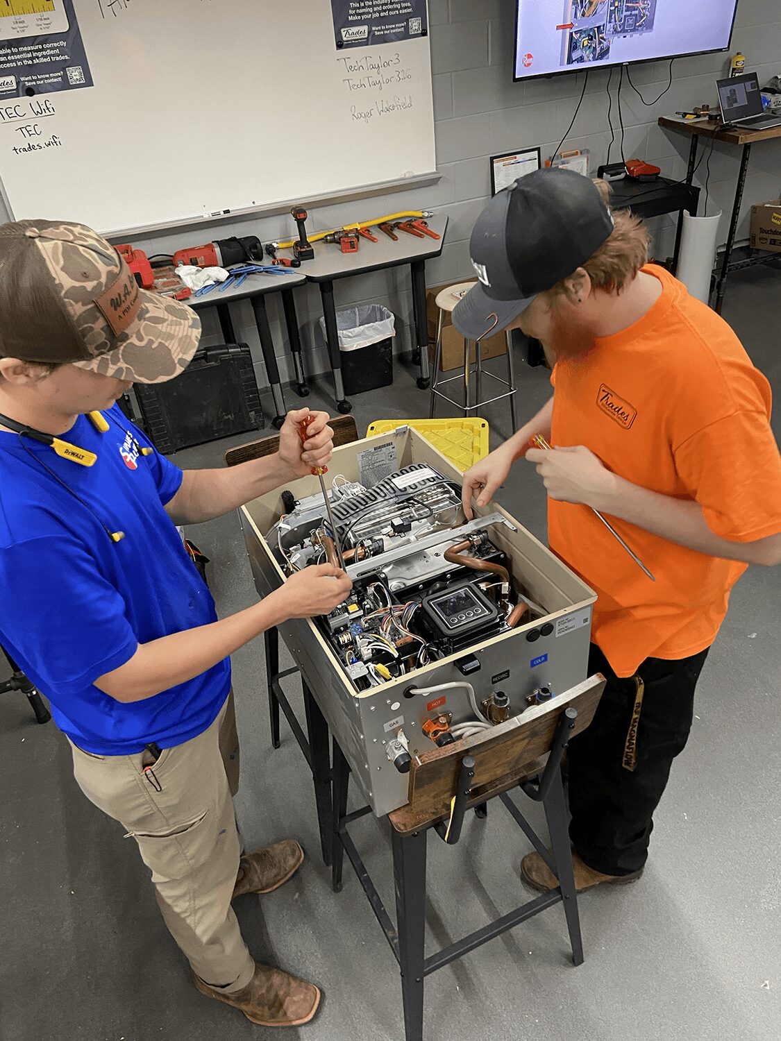 Two boys working on an open electronic device on a table in a classroom, with a whiteboard and monitor in background.