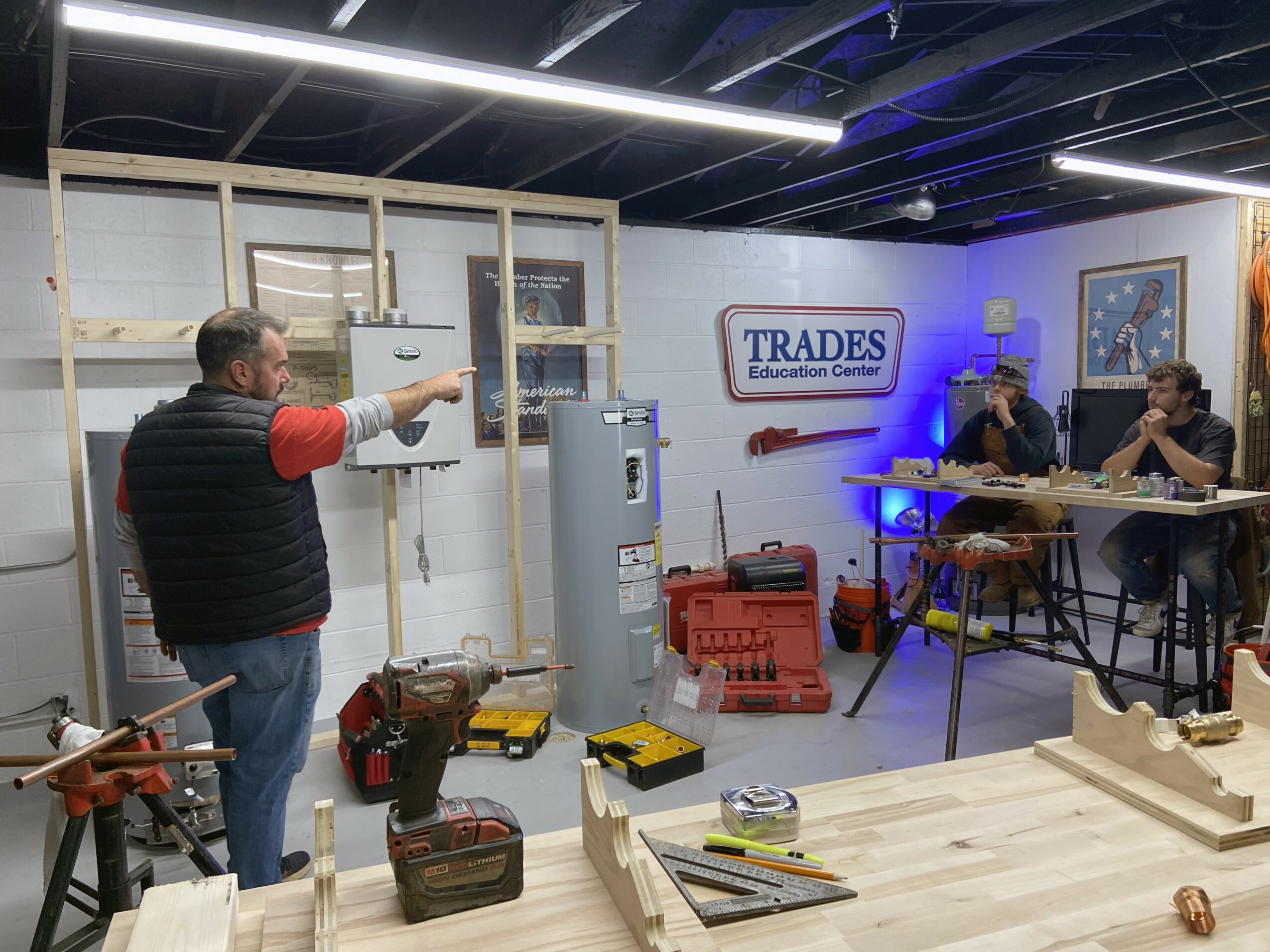 Person in a workshop pointing at a wall with tools and equipment, with two people working at a table in the background.
