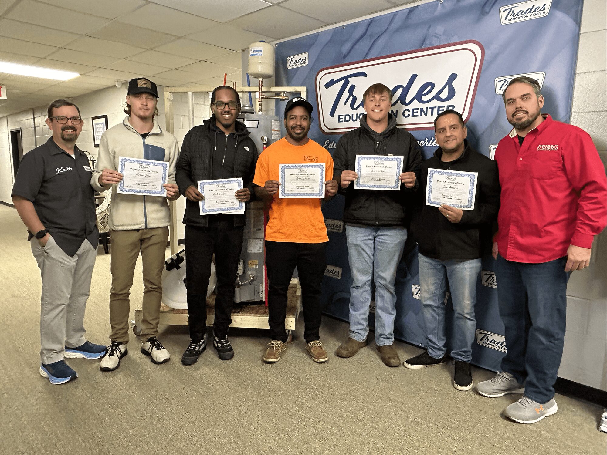 Group of seven people standing indoors, holding certificates, smiling, in front of a banner and equipment.