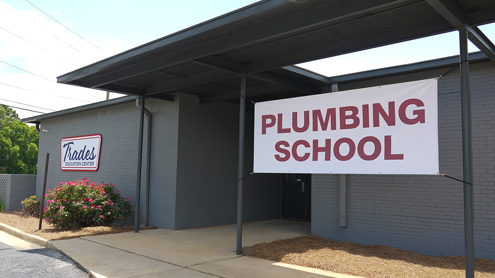 Building with a sign reading 'PLUMBING SCHOOL' and a smaller sign that says 'Trades' near some bushes.
