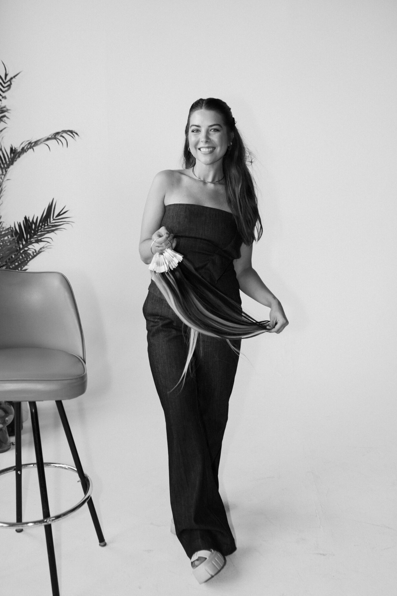 Woman in black dress holding a fabric, standing next to a chair and a plant, smiling at the camera.