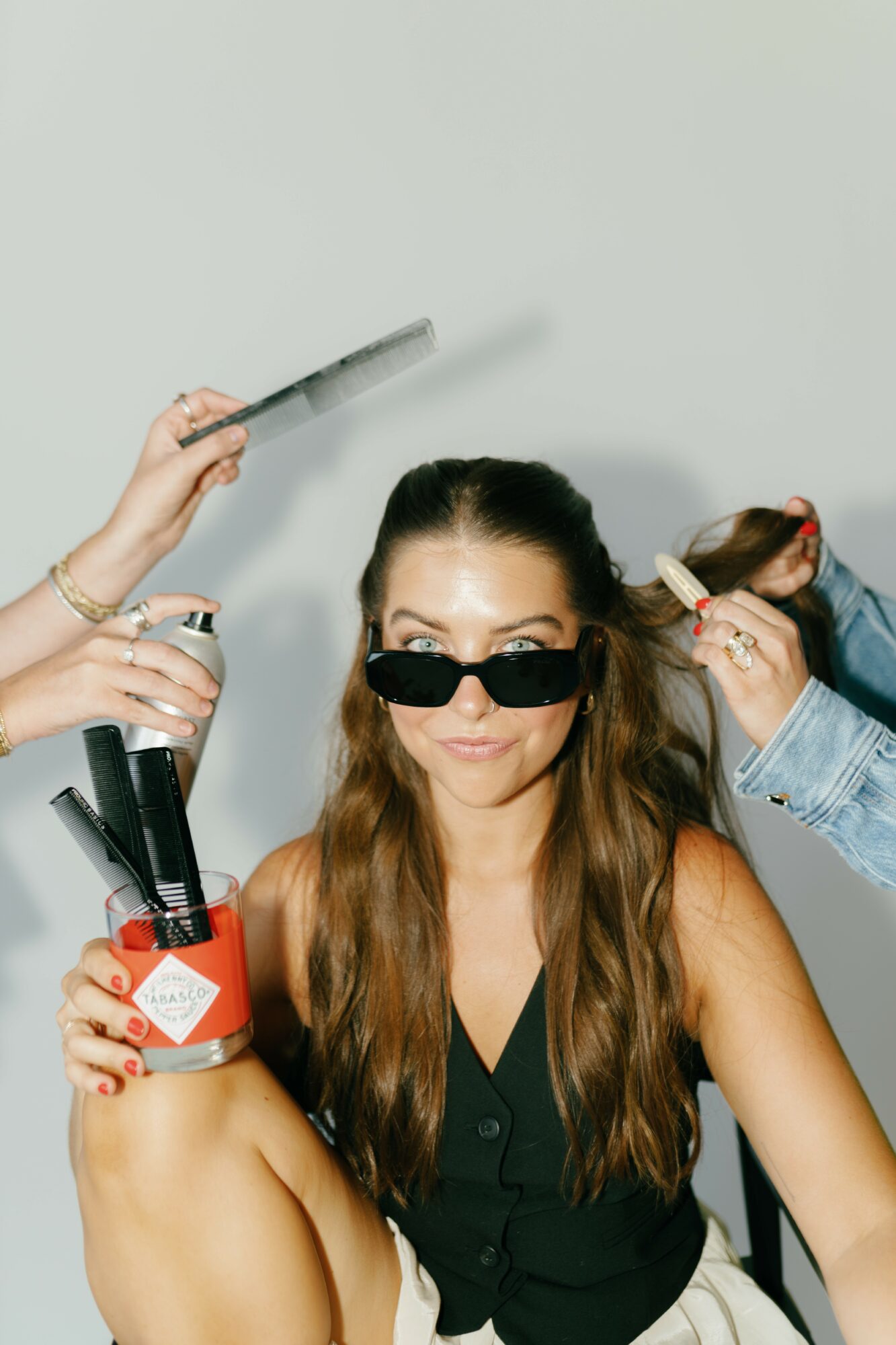 Woman with long hair wearing sunglasses, surrounded by hands holding makeup brushes and tools, against a plain background.