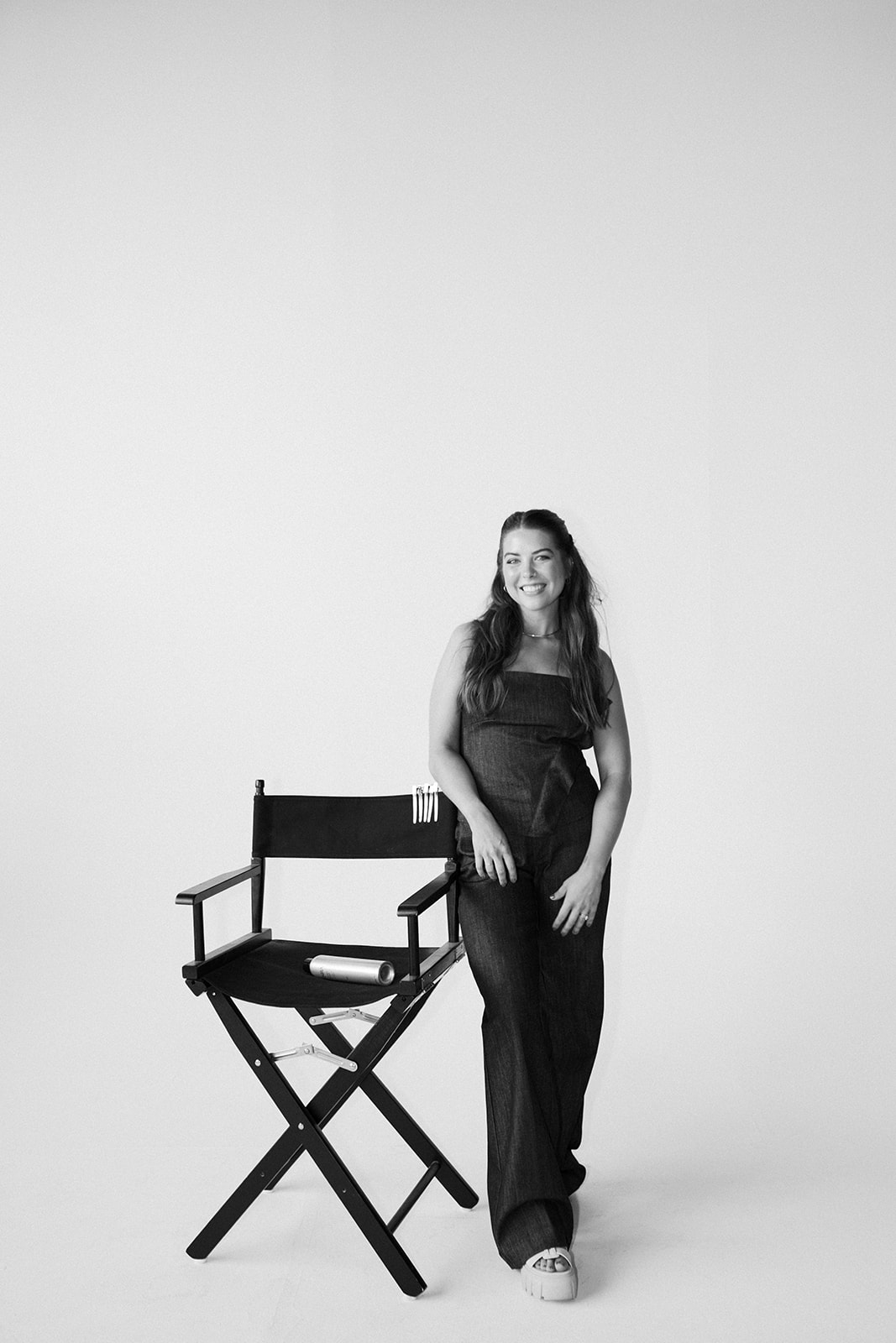Woman standing next to a director's chair, smiling, in front of a plain background.