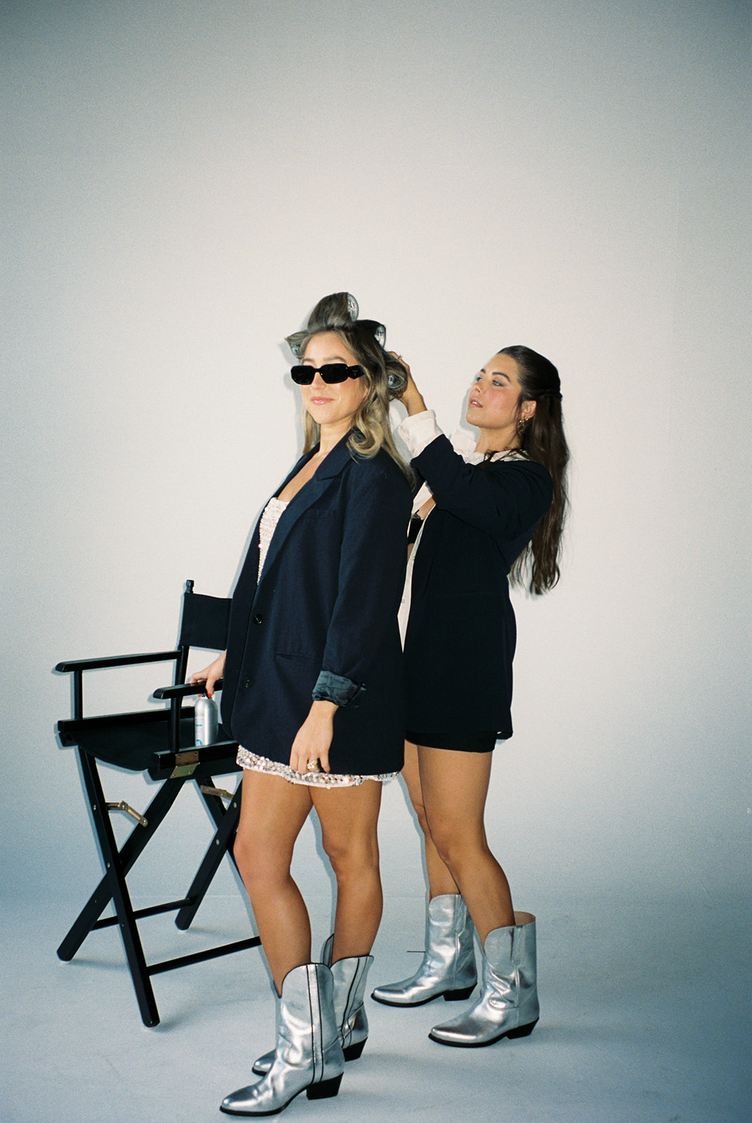 Two women in black outfits and silver boots, one adjusting sunglasses, standing next to a director's chair against a plain background.