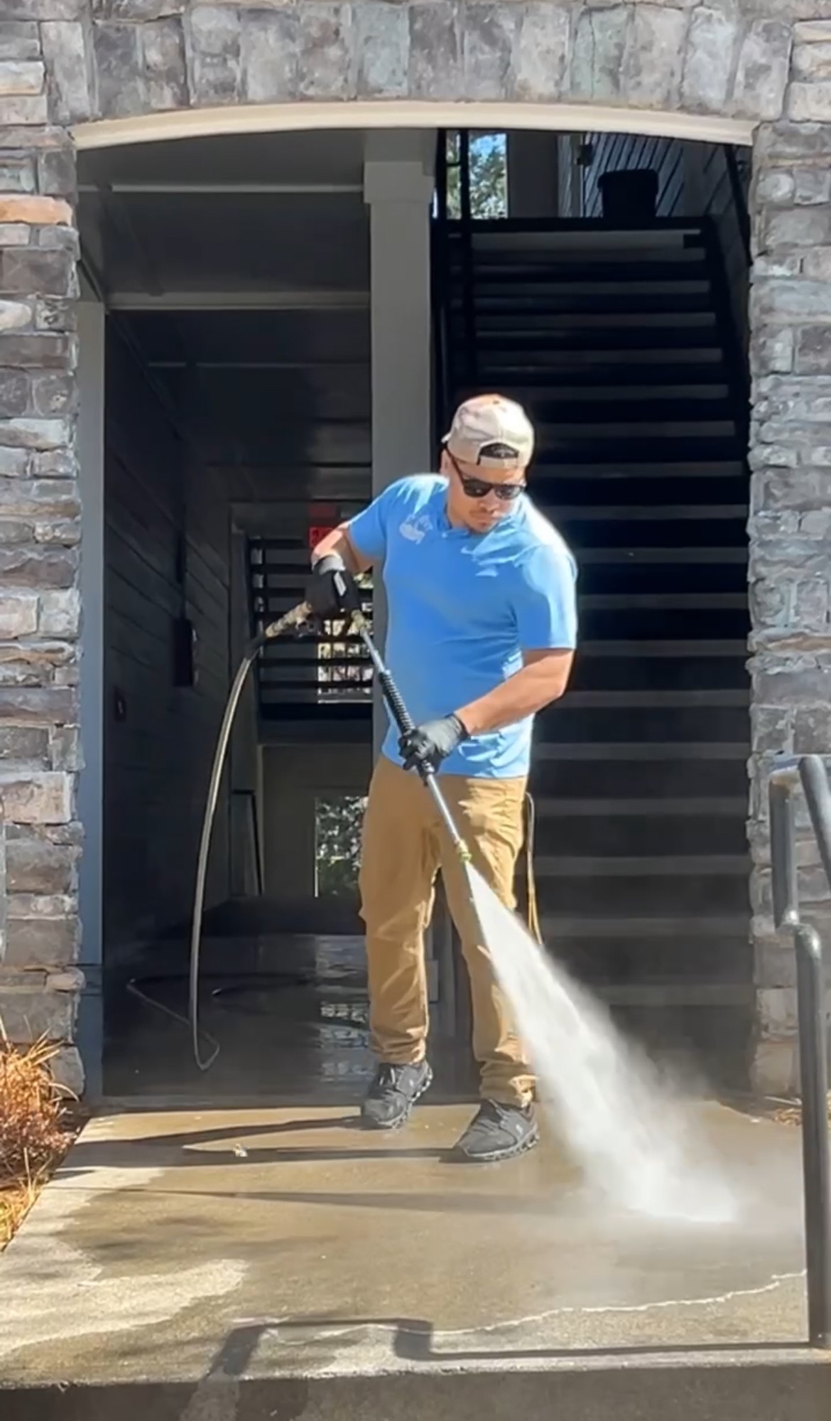 Person in blue shirt and beige pants using a pressure washer on a concrete surface outside a building.