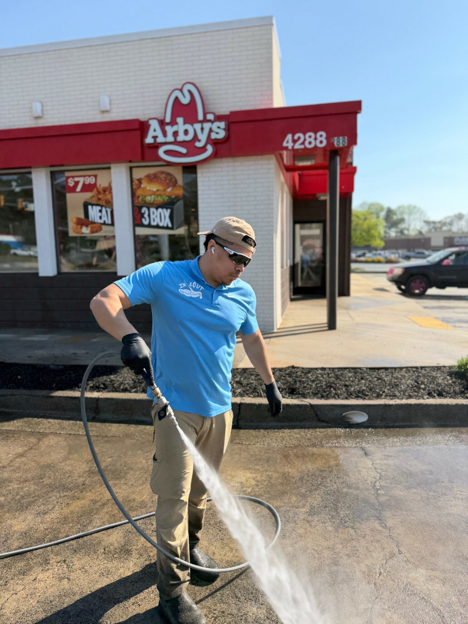 Person cleaning outside Arby's restaurant with a high-pressure water hose, wearing sunglasses, a blue shirt, and gloves.