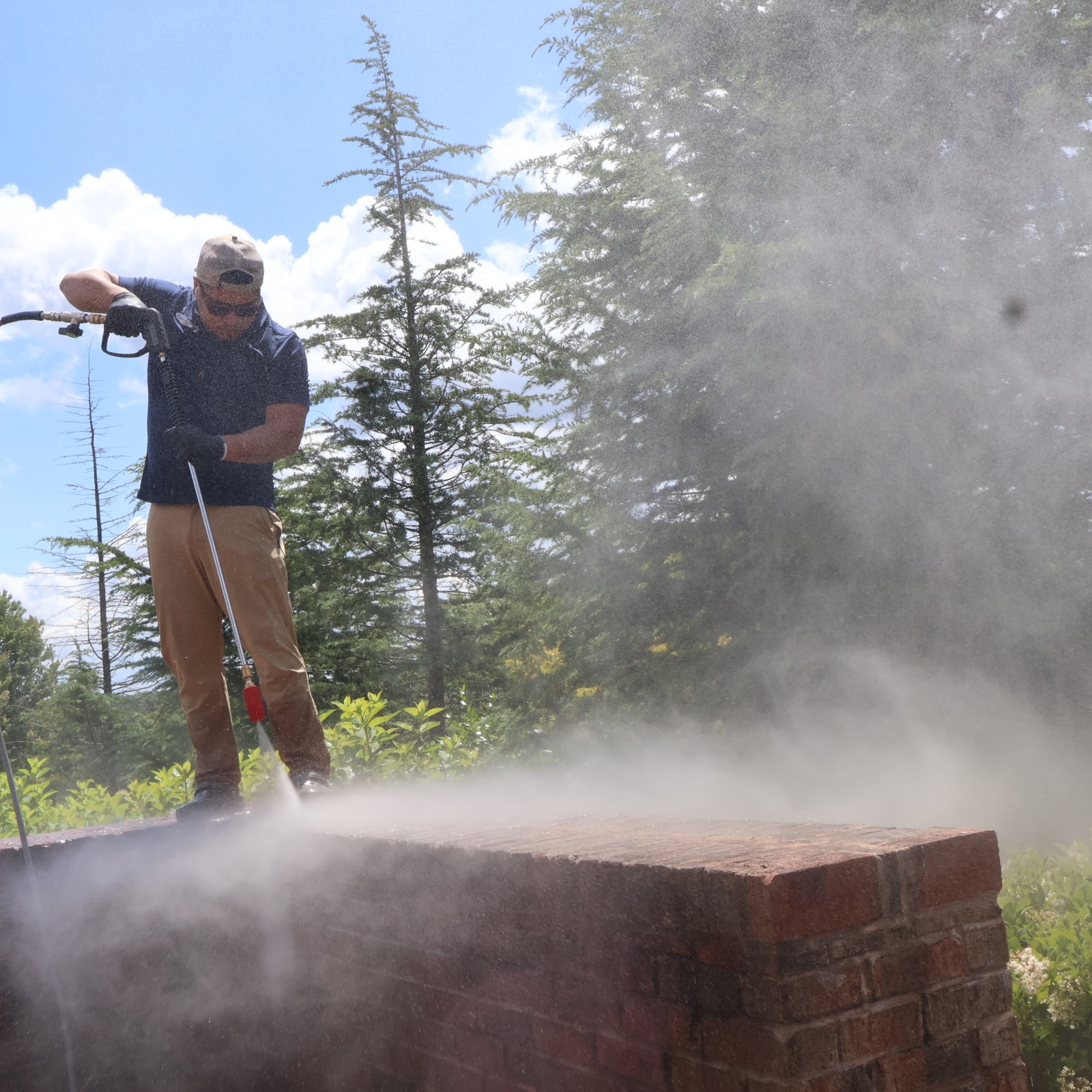 Person in outdoor setting using a tool, with trees and sky in background, and dust or smoke near a brick structure.