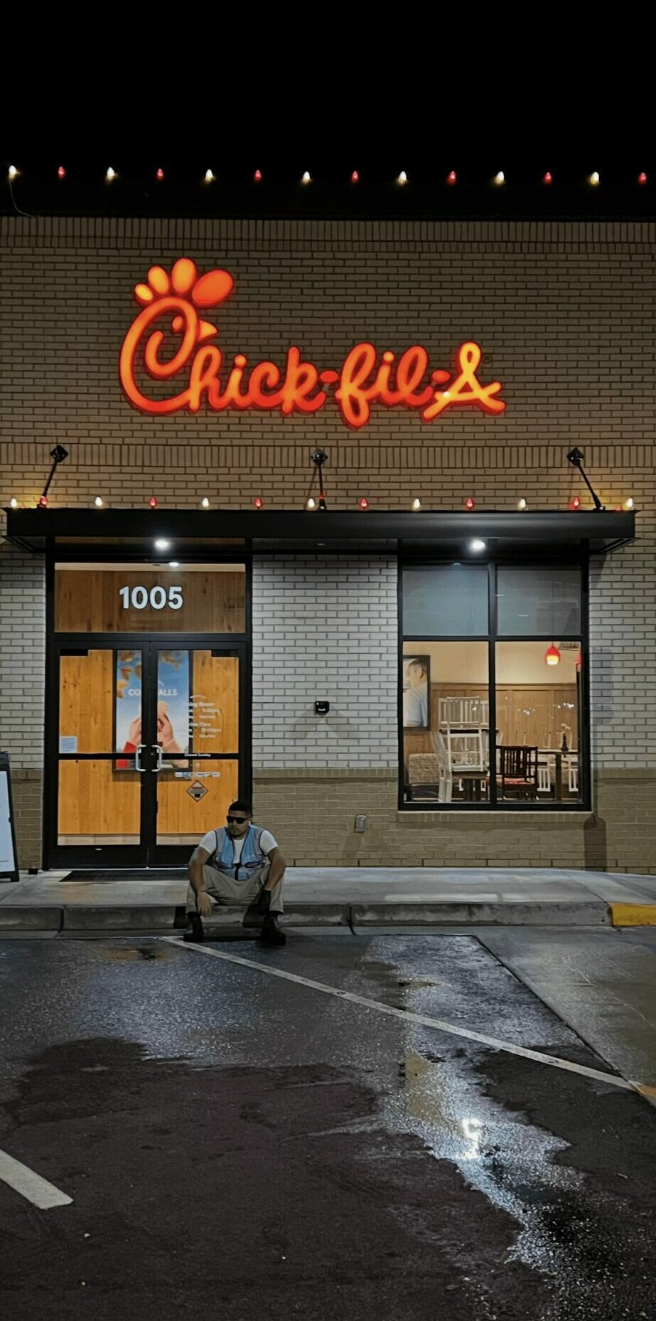 Chick-fil-A sign above the restaurant entrance at night, with a person sitting on the sidewalk in front.