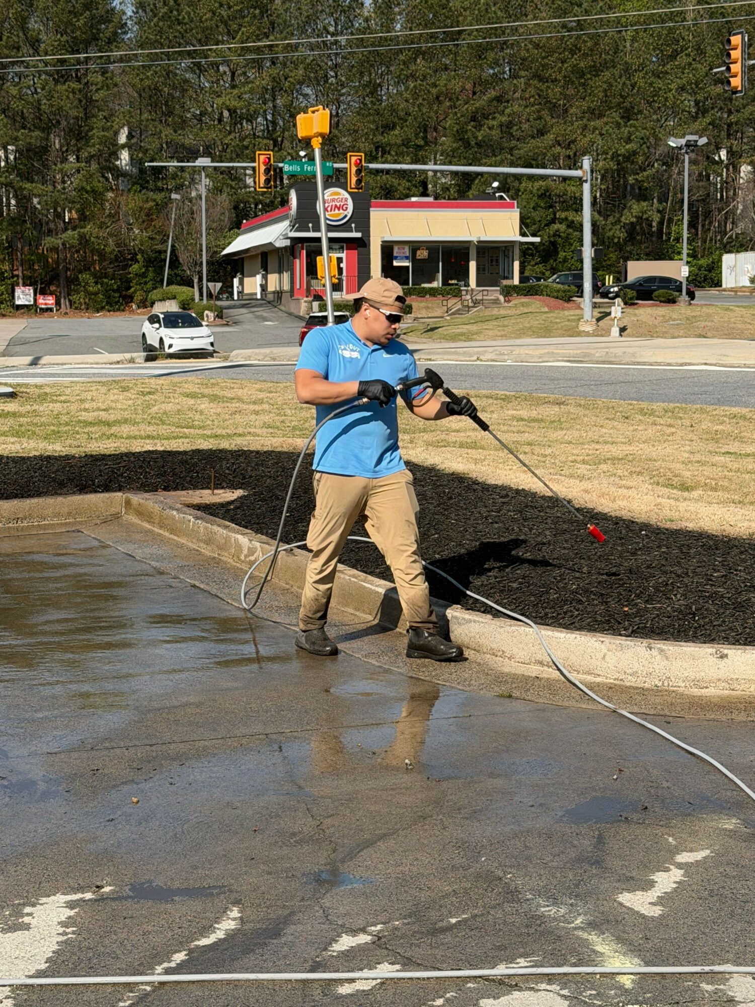 Person using a power washer on a concrete surface outdoors, wearing a blue shirt, beige pants, and sunglasses.