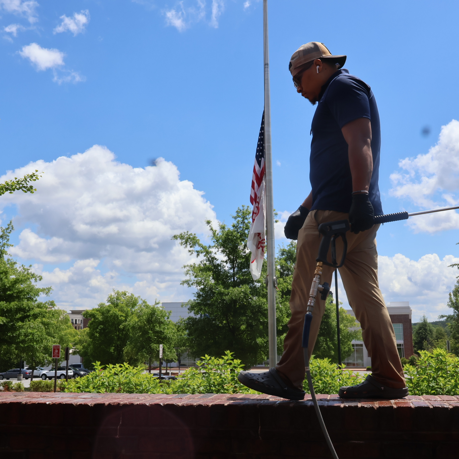 Man in dark shirt and khaki pants holding a metal detector on a brick ledge outdoors with trees and a flagpole nearby.