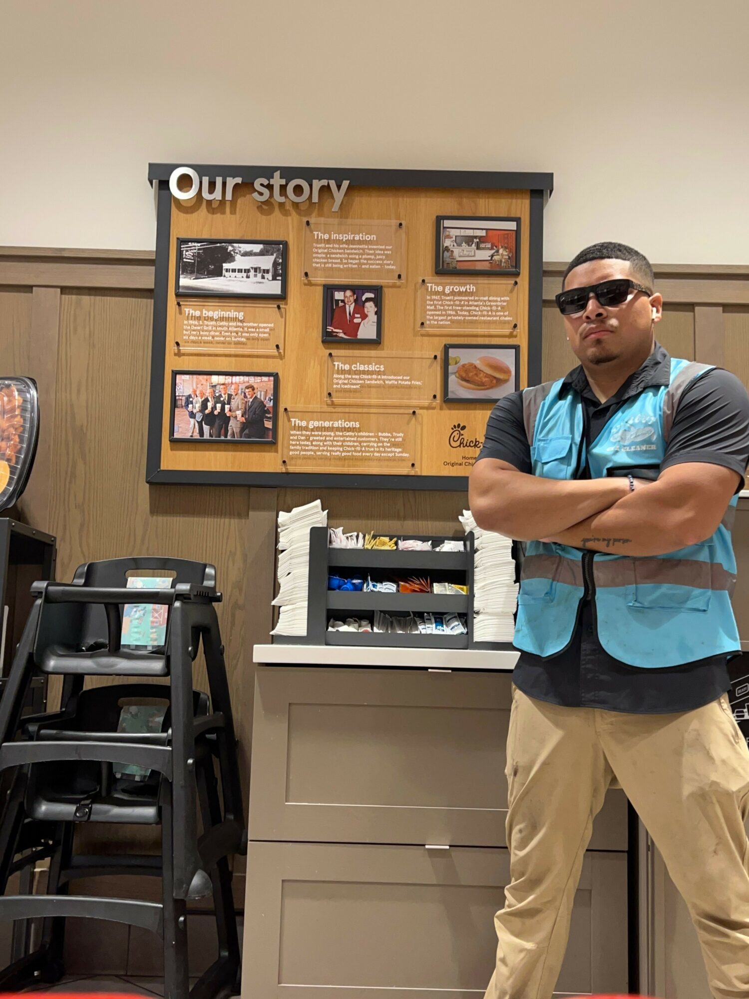 Man wearing sunglasses, blue vest, black shirt, and beige pants standing with arms crossed in front of a display board and counter.