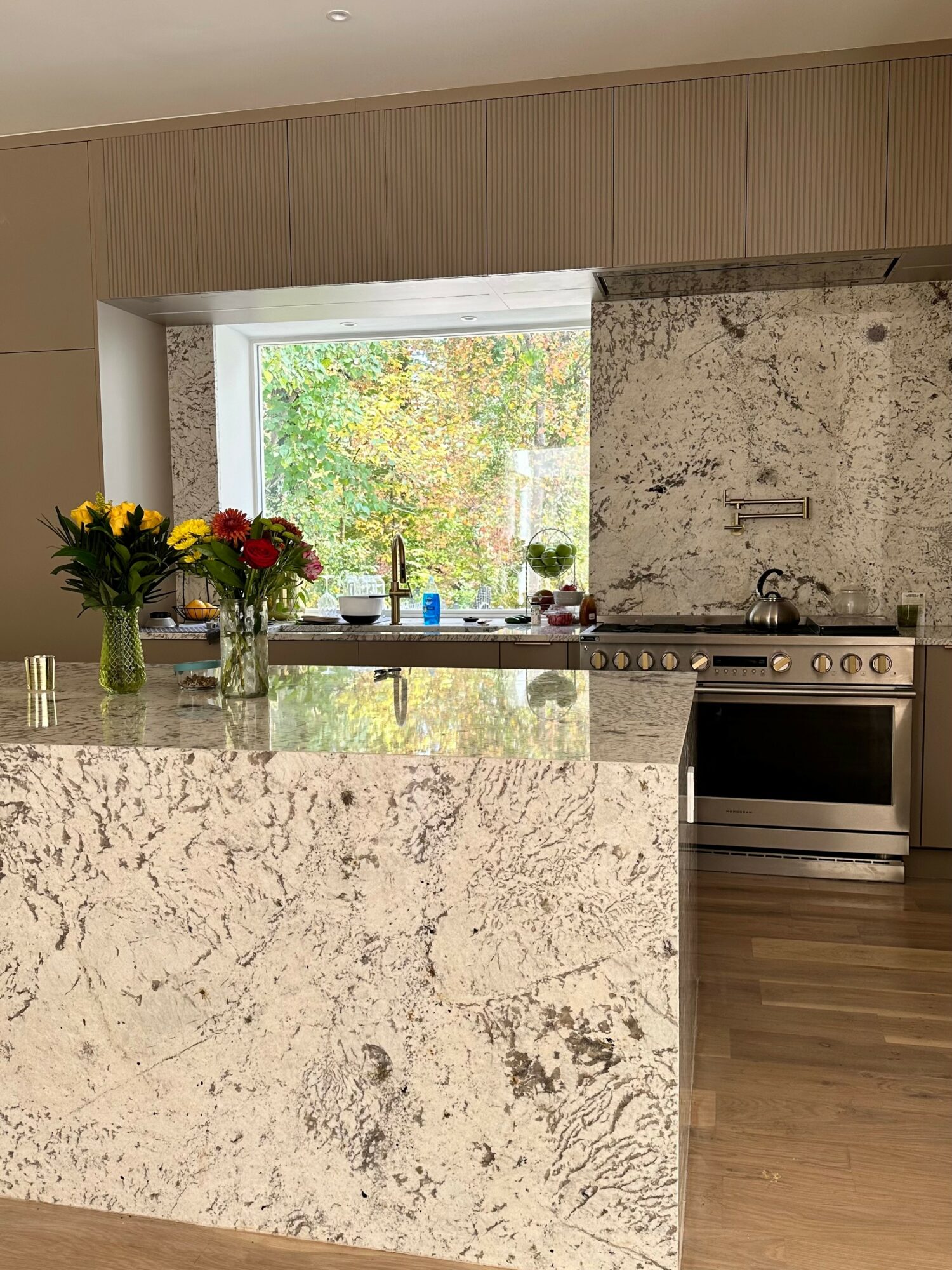 Kitchen with marble island, flowers in vases, stove, and window showing outdoor trees.