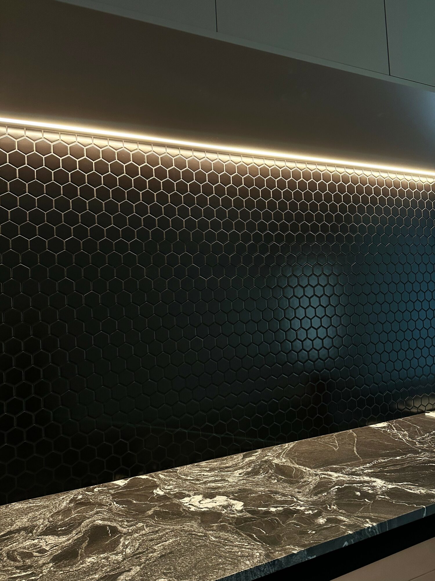 Close-up of a black hexagonal tile wall with a strip of LED lighting above a marble countertop.