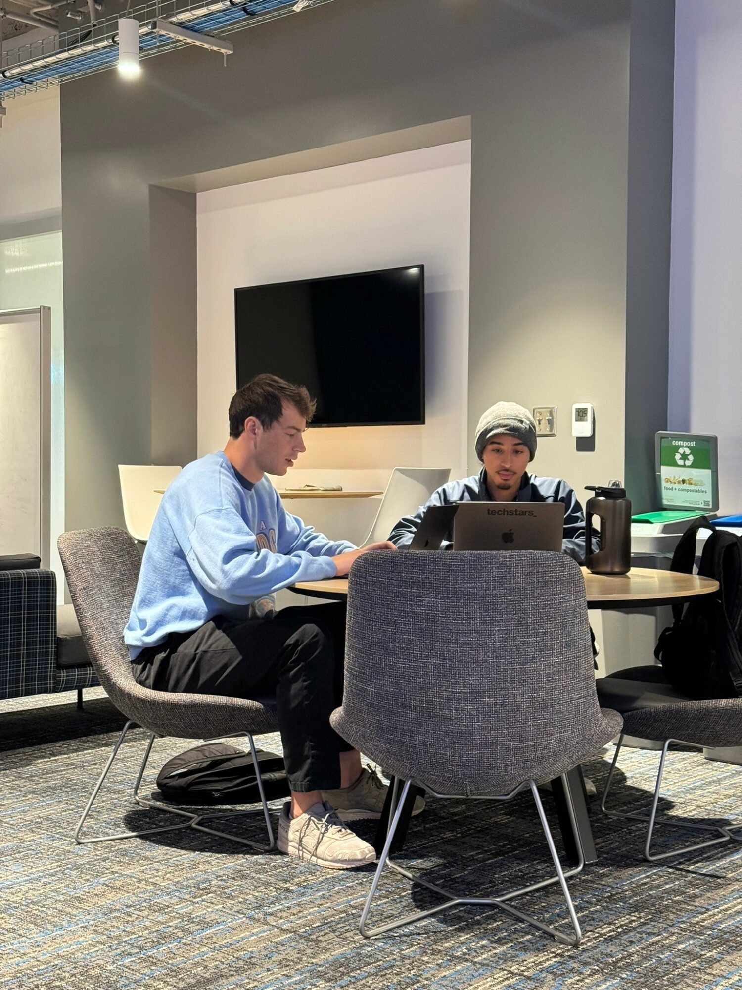 Two men sitting at a round table with laptops in a modern office space, with a large TV on the wall behind them.