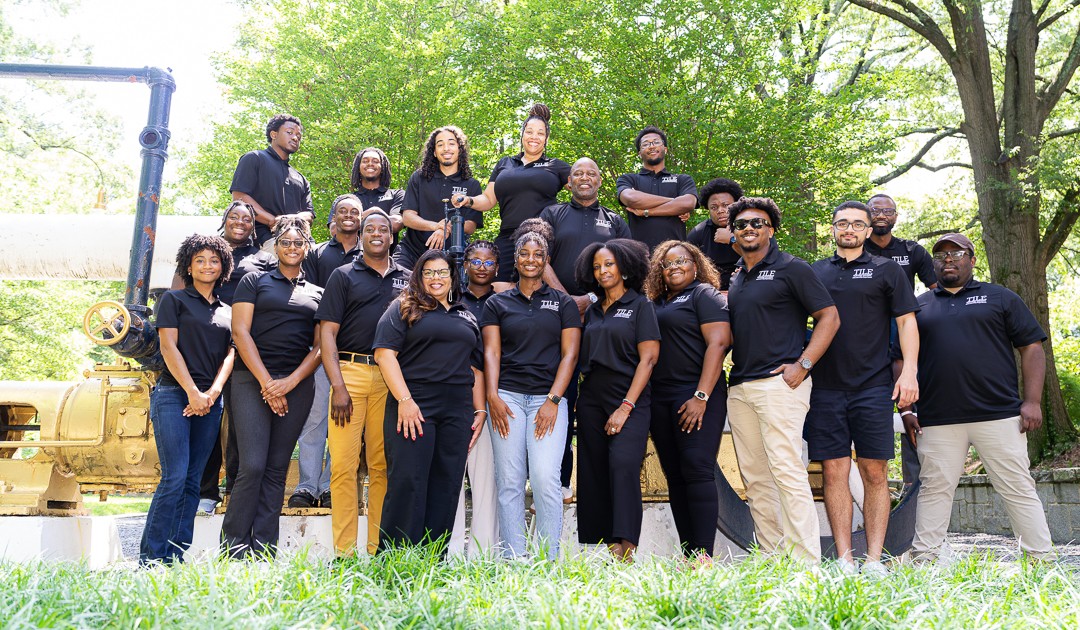 Group of people standing outdoors on grass with trees in background, posing for a photo, wearing dark shirts.