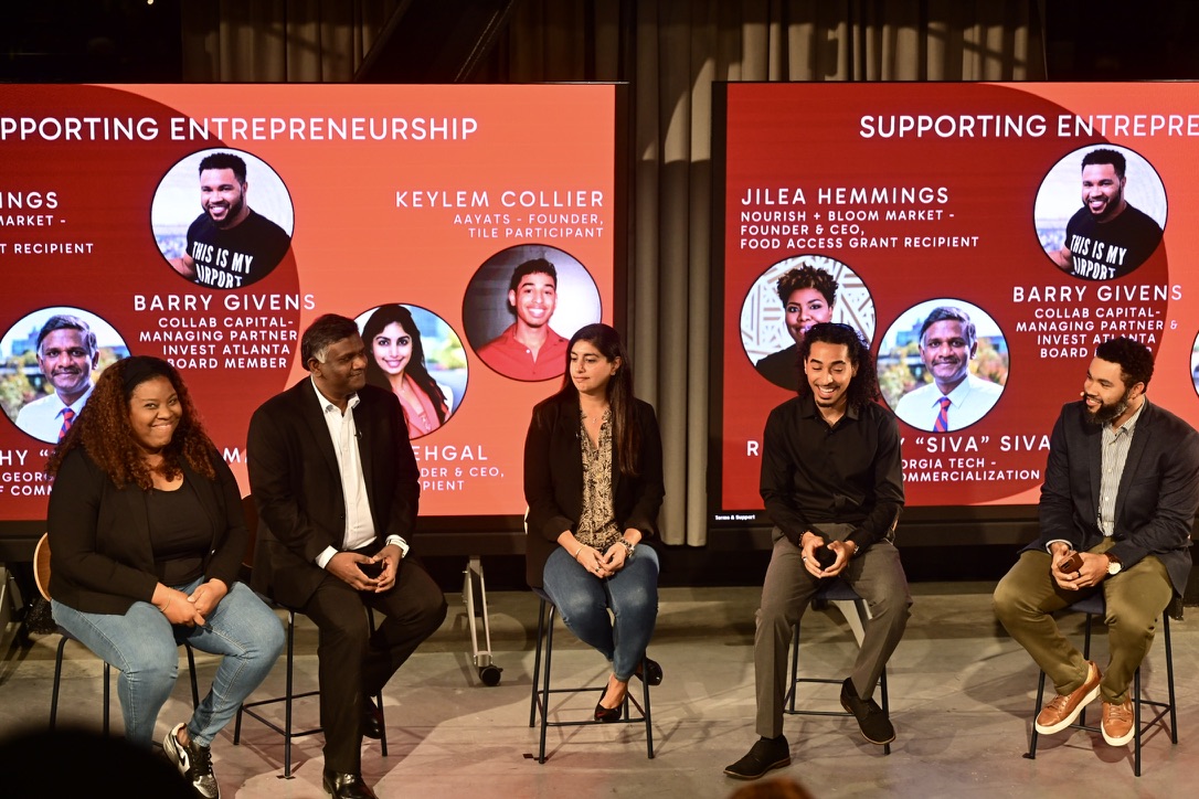 Six people seated on stage in front of red banners with text and images, participating in a panel discussion.