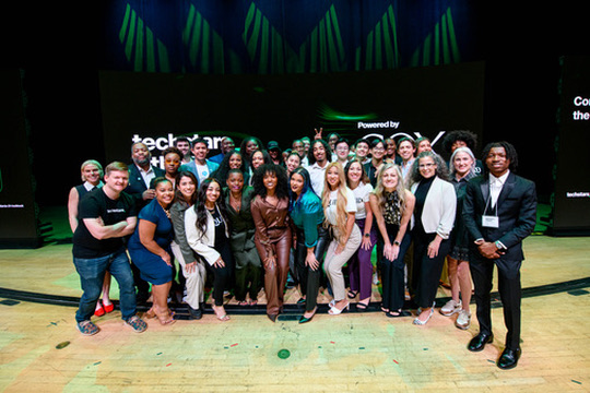 Group of people on stage in front of a black screen, some smiling and posing for a photo.