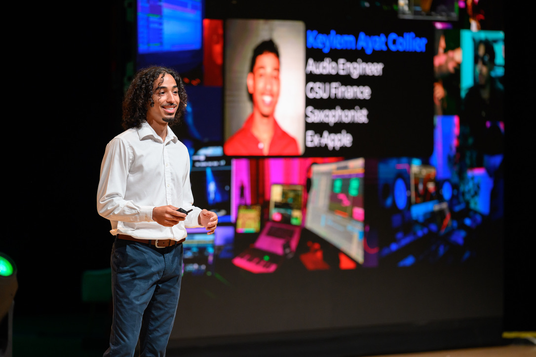Woman speaking on stage with a large screen displaying her photo and text behind her.
