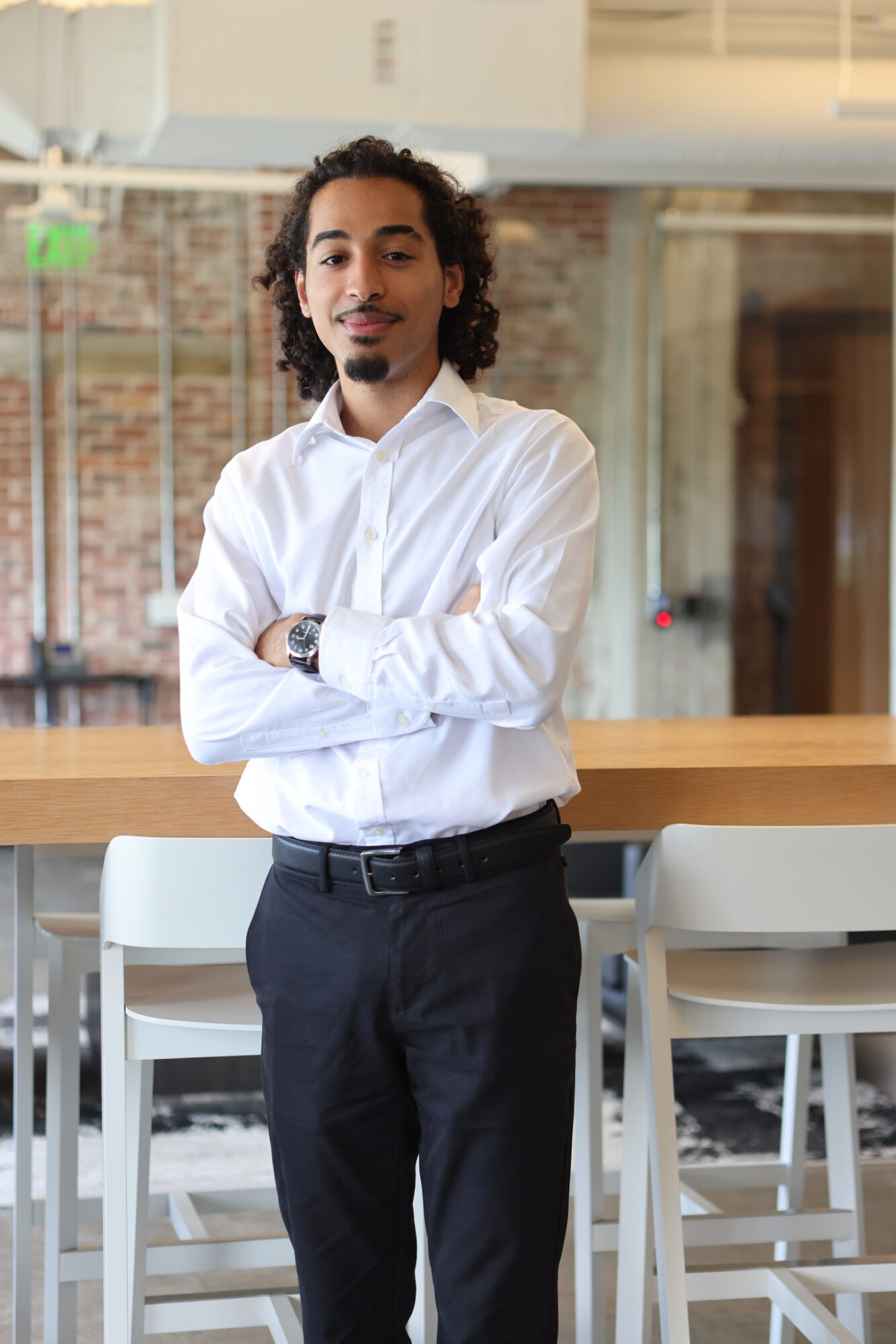 Man with curly hair and a goatee, wearing a white shirt and dark pants, standing with arms crossed in a modern office.