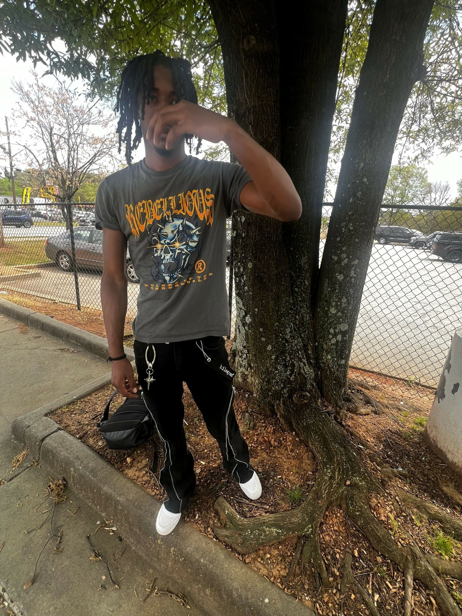 Person standing outdoors near a tree, wearing a graphic t-shirt, black pants, and white shoes, holding a phone.