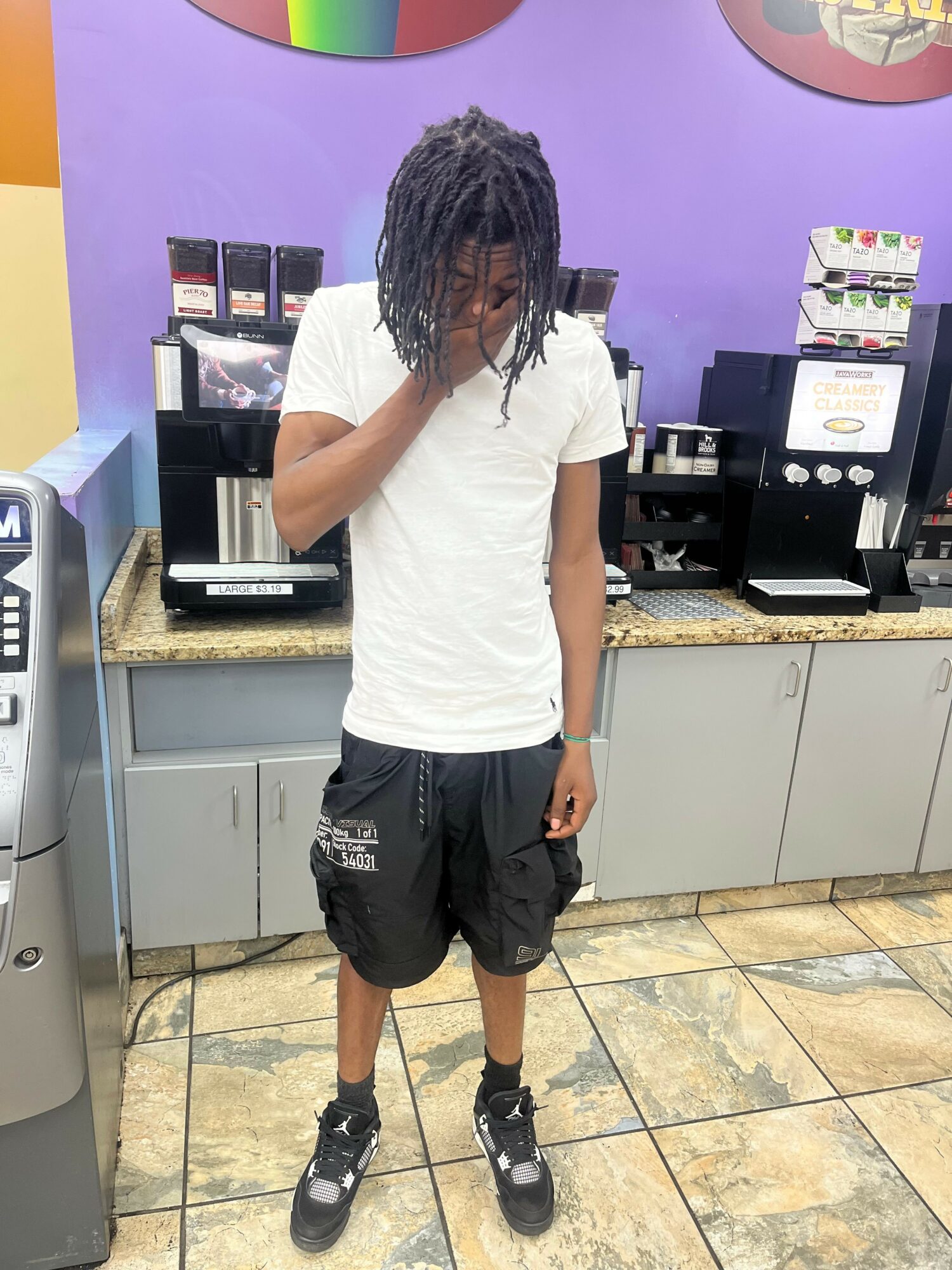 Young man with dreadlocks in a convenience store, wearing a white t-shirt and black shorts, looking down.