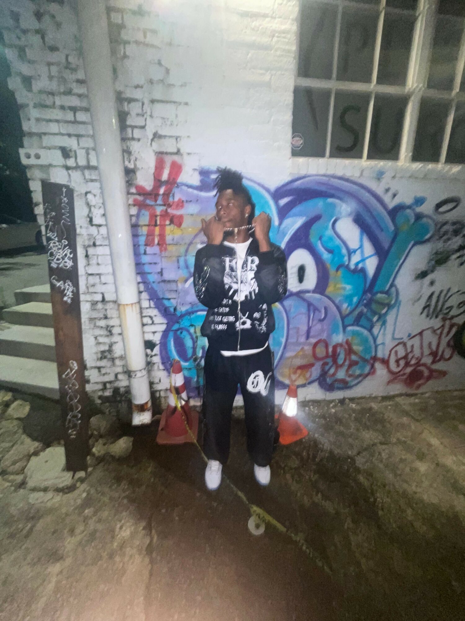 Person standing in front of graffiti wall, holding a phone, wearing black and white clothing, with a building window nearby.