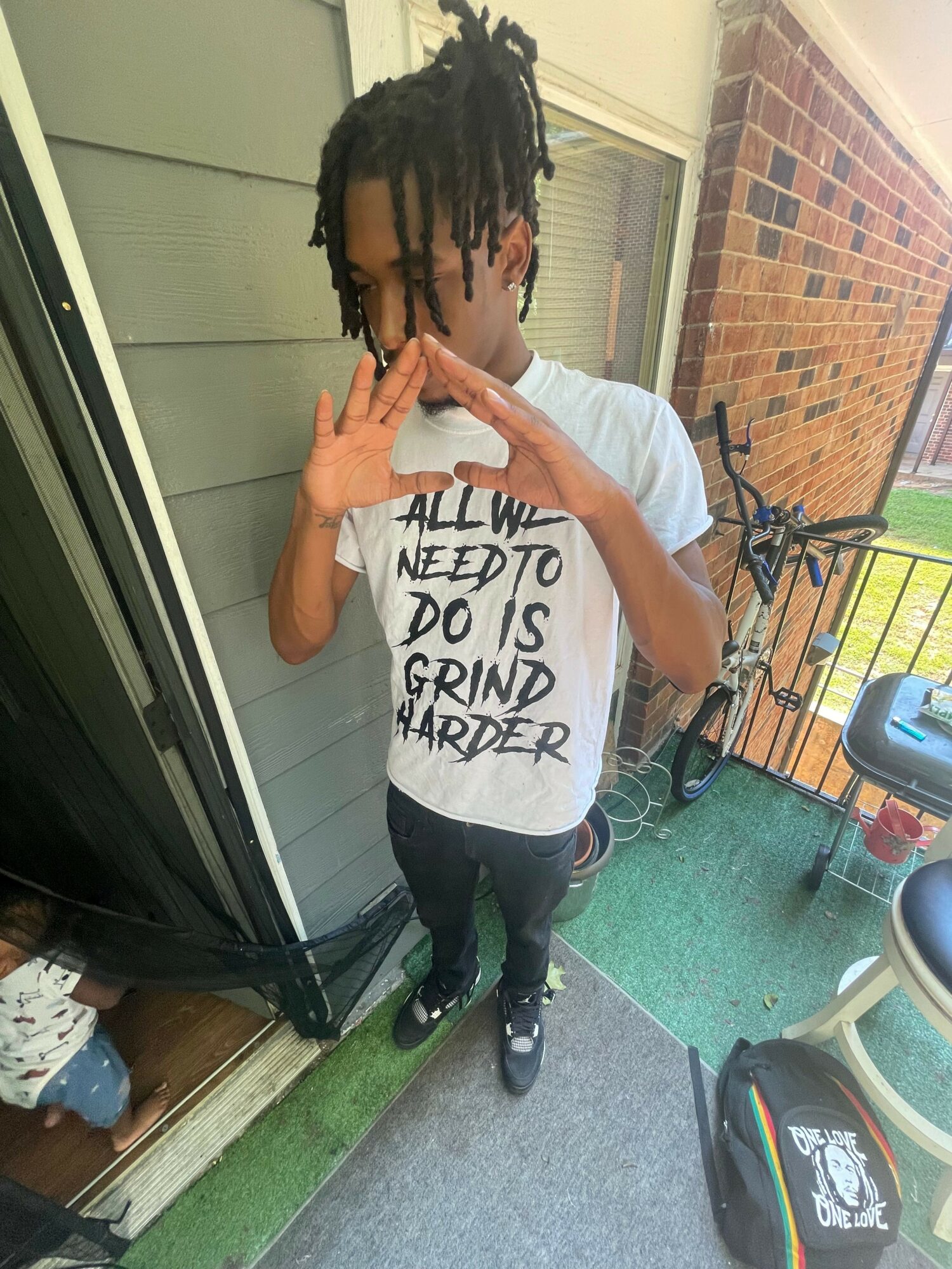 Young person with dreadlocks taking a selfie outside, wearing a white T-shirt with text, standing on a porch with a brick wall and railing.