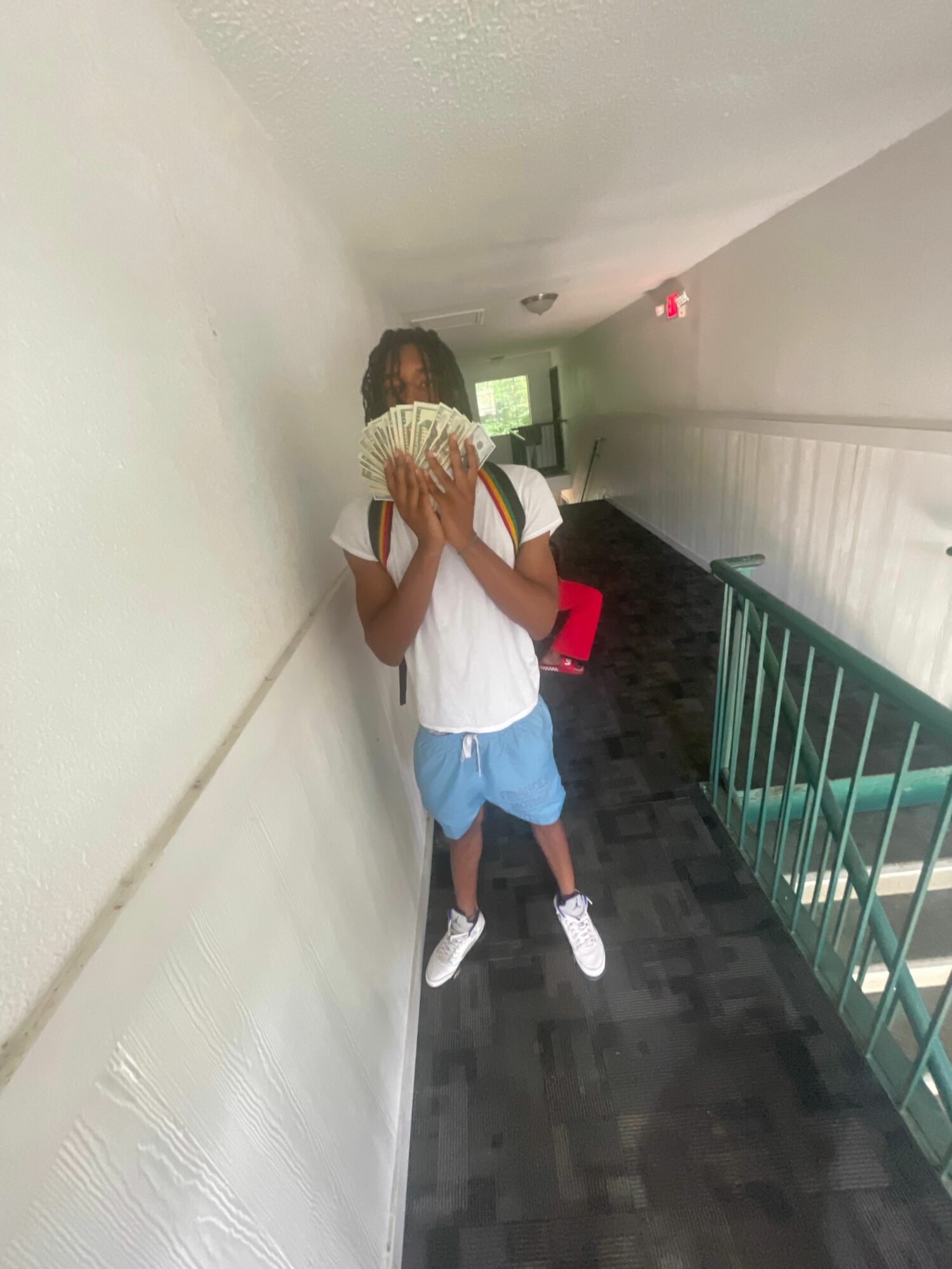 Person standing in hallway holding a fan covering face, wearing white shirt, blue shorts, and white shoes.