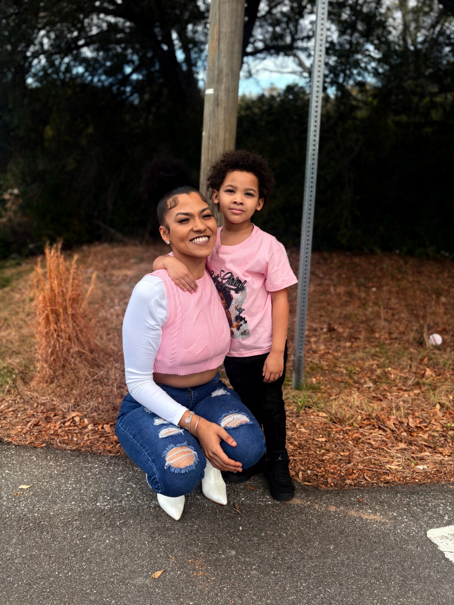 Two smiling girls standing outdoors near a pole and trees, one kneeling and the other standing, both wearing pink shirts.