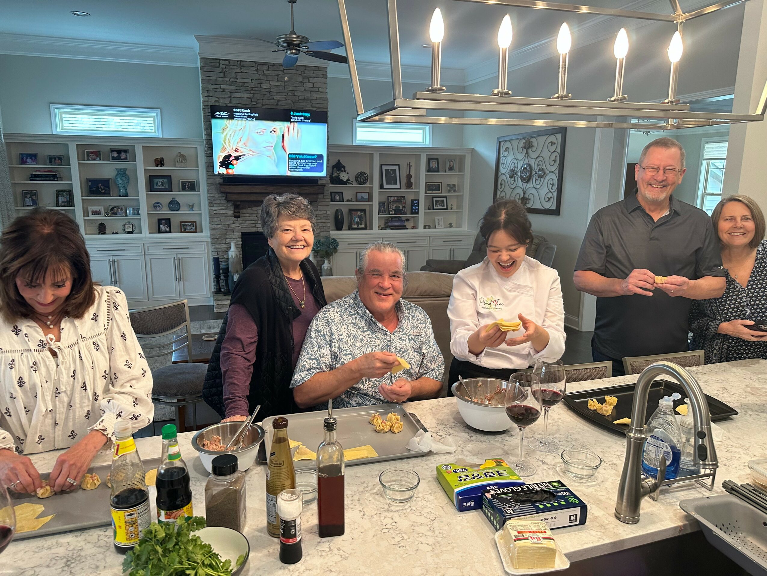 Group of six people gathered around a kitchen island, smiling and preparing food together indoors.