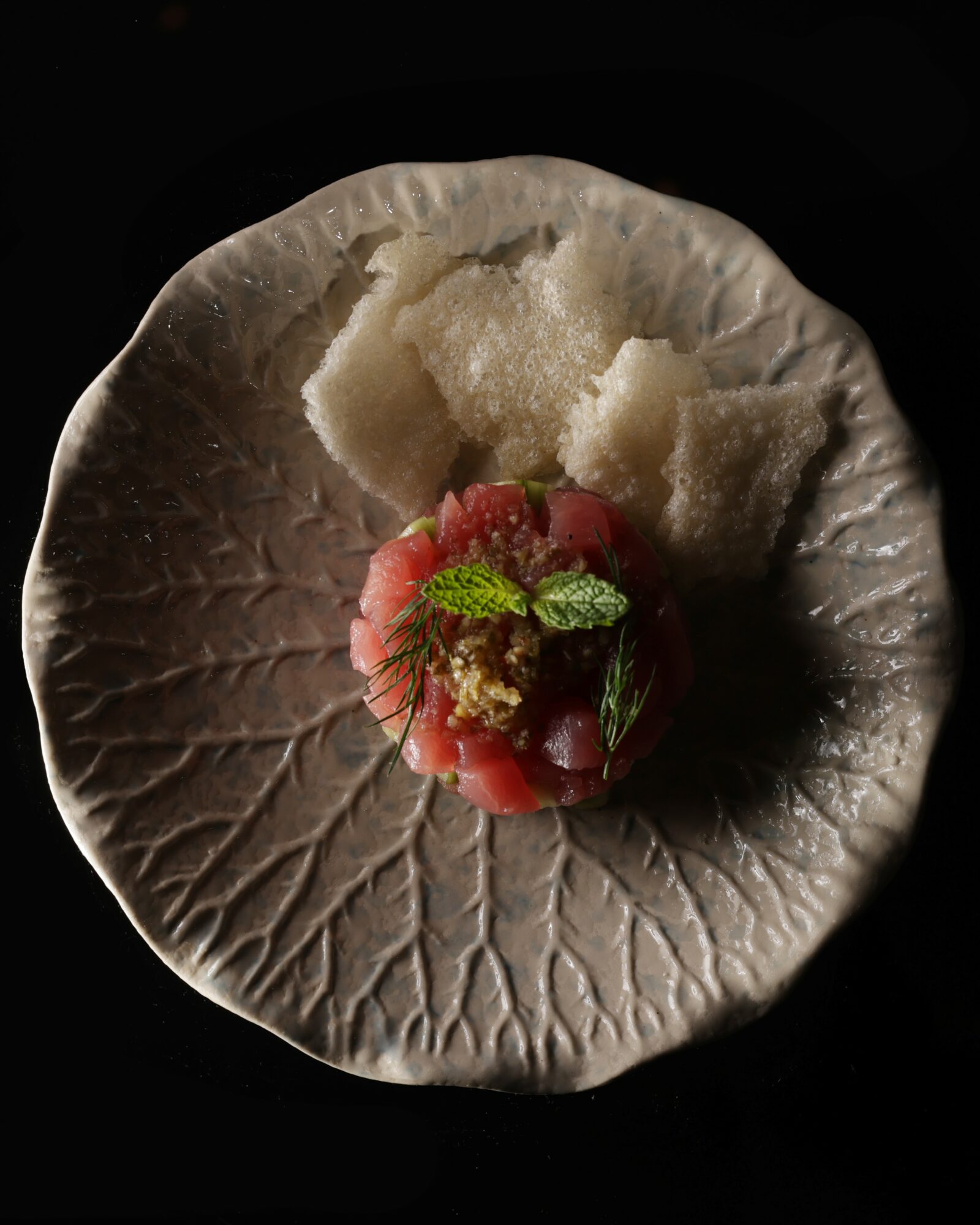 A round, textured plate with a small portion of food, garnished with herbs, on a black background.