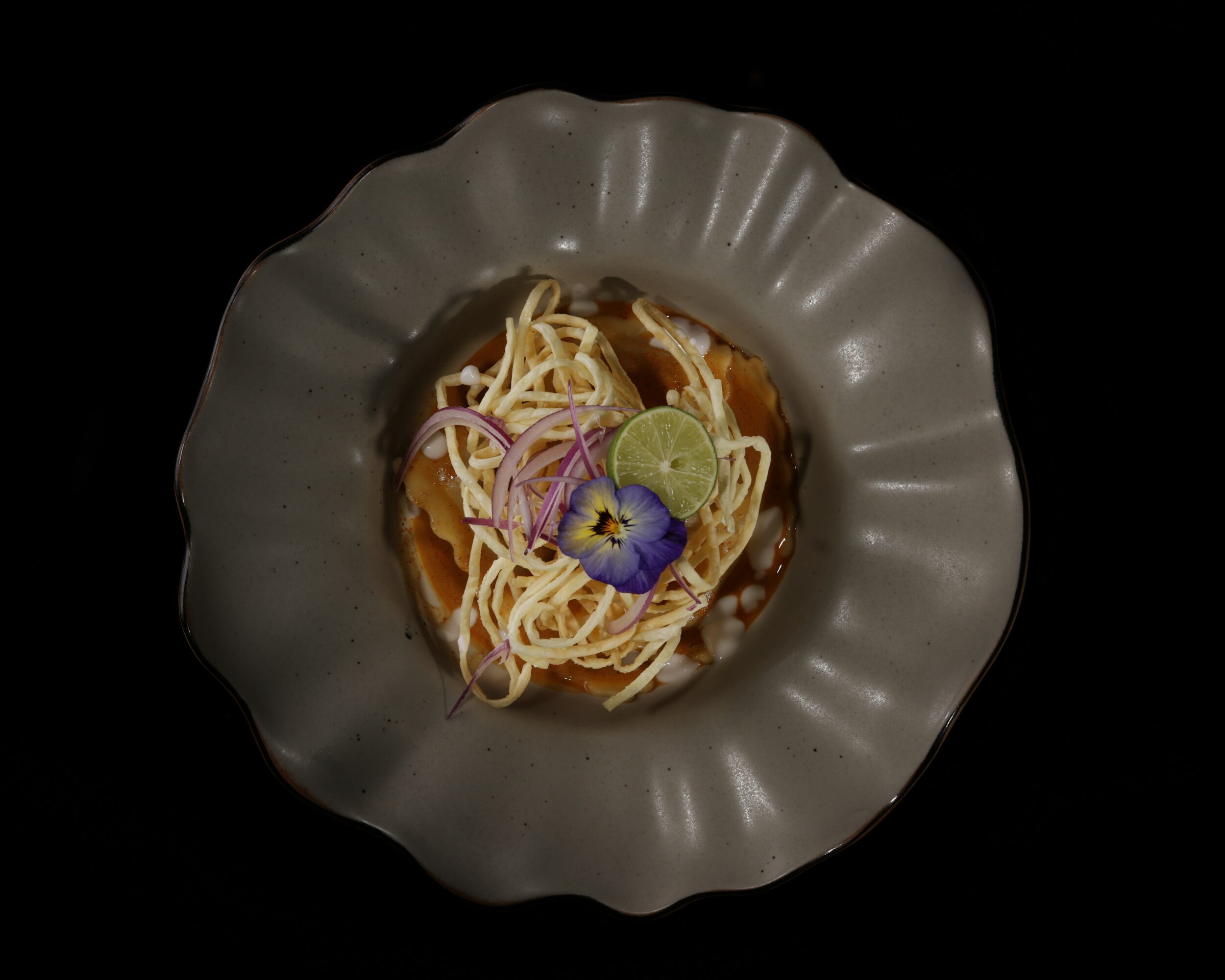Bowl of noodles with lime, onion, and edible flower garnish on black background.