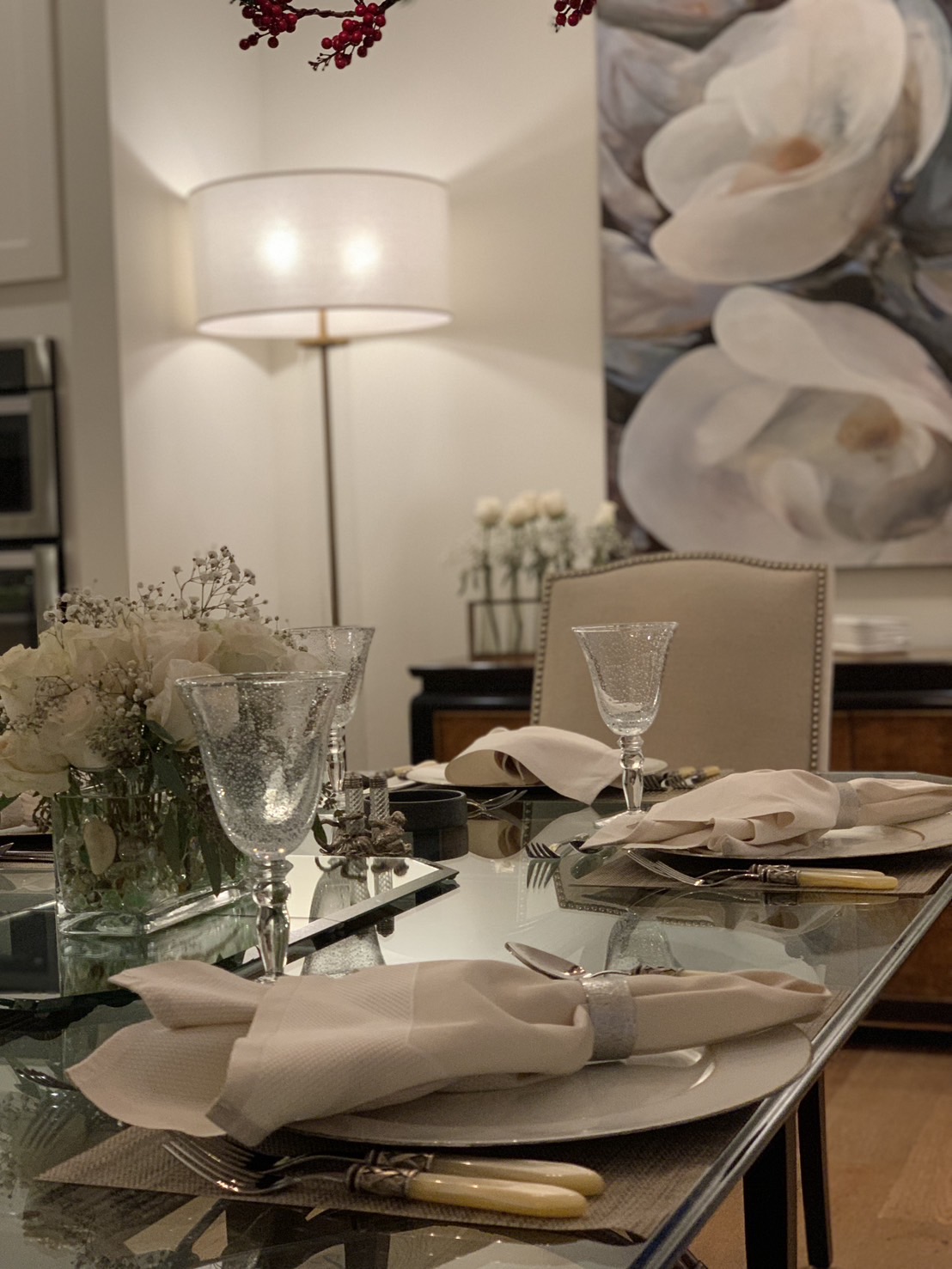 Dining table set with glasses, plates, napkins, and cutlery, with a floral arrangement and a painting in the background.