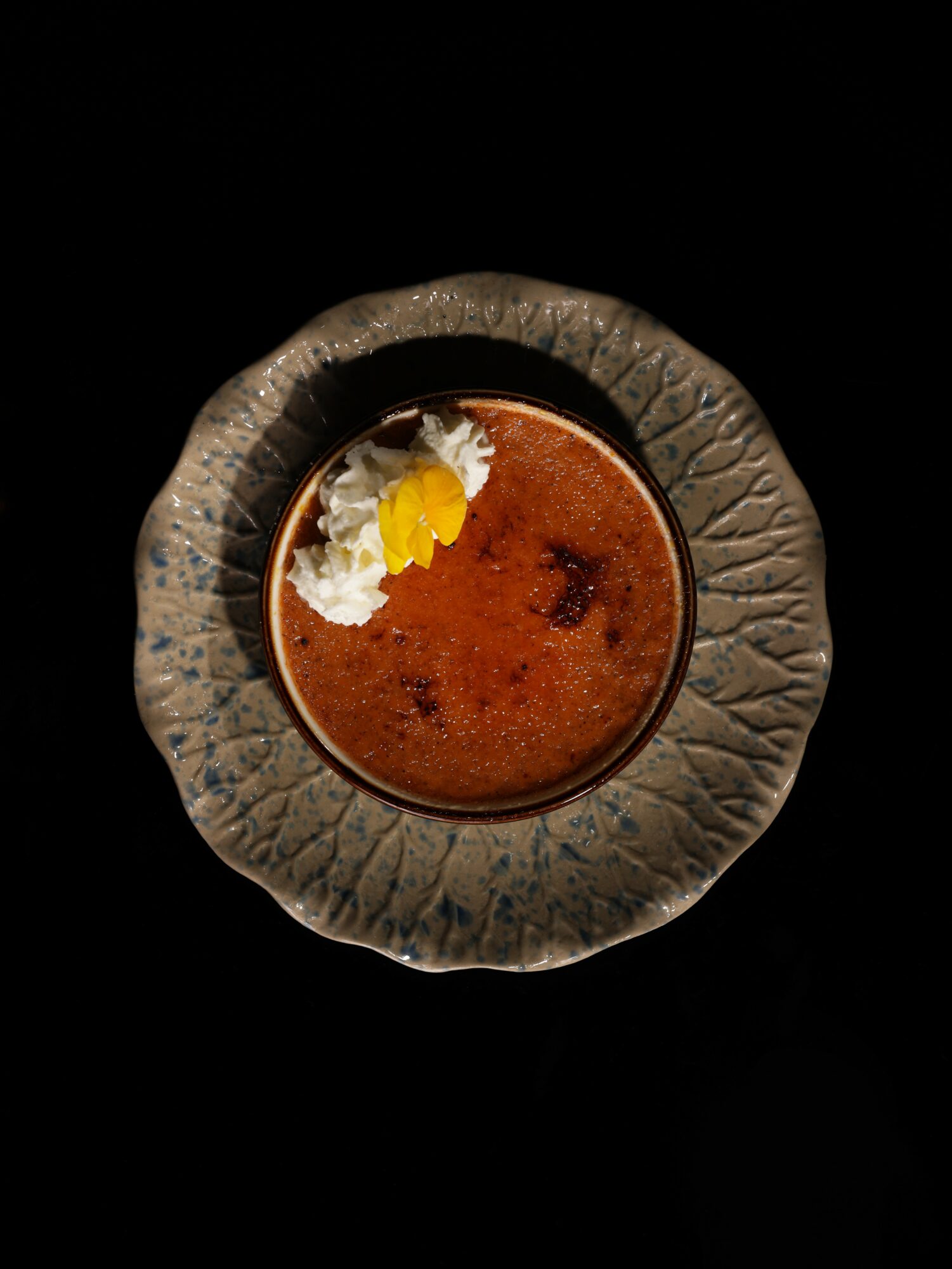 Top view of a bowl with a brown dessert garnished with white cream and a yellow flower petal, on a textured gray plate.
