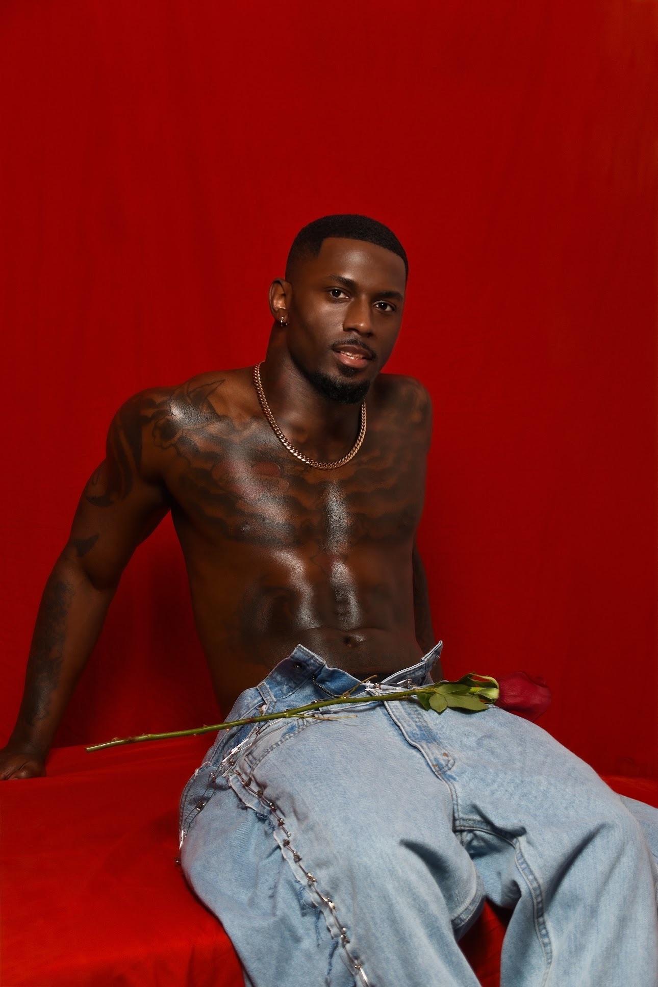 Young man with tattoos sitting shirtless against a red background, wearing jeans and holding a flower.