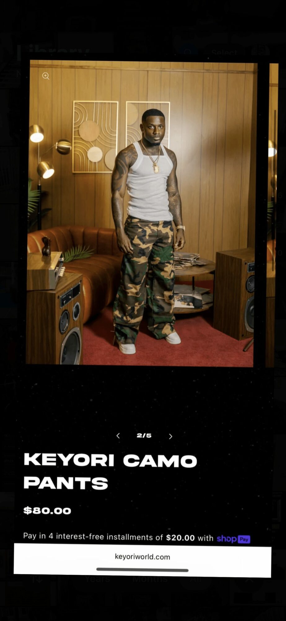 Man standing in a room with wooden walls, wearing a white tank top and camouflage pants, surrounded by speakers and lamps.