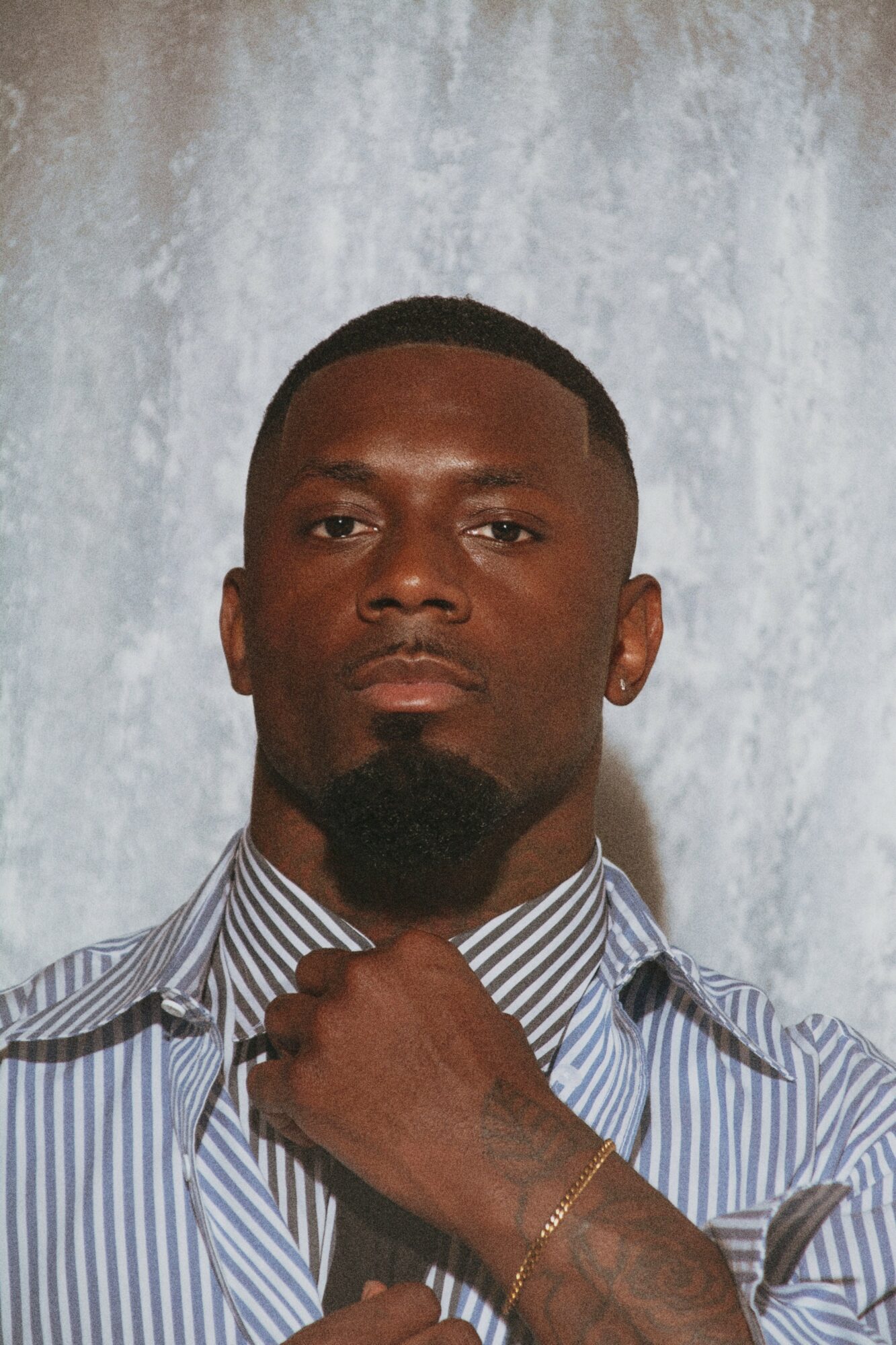 Man with short hair and beard wearing a striped shirt, holding his collar, standing in front of a textured background.