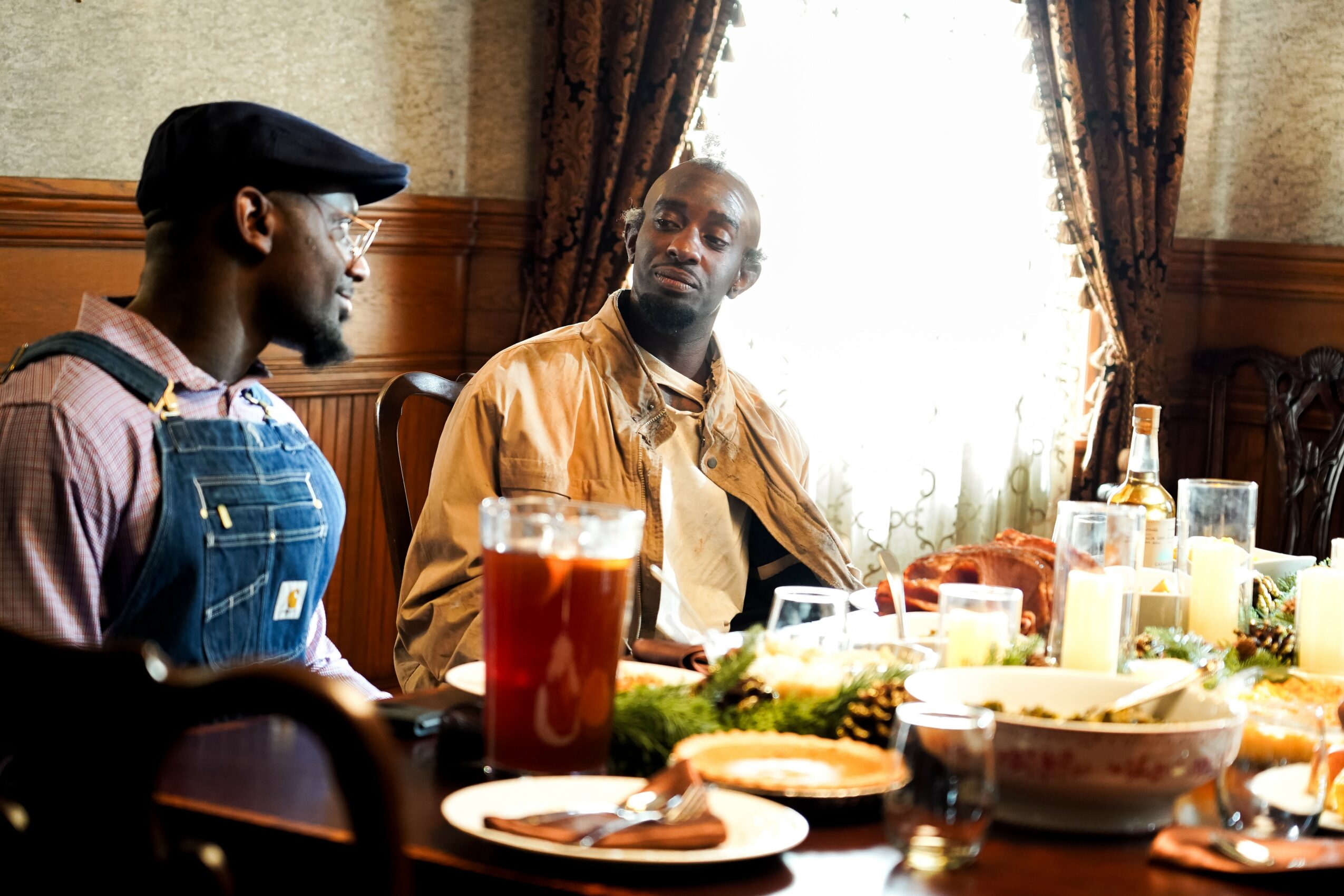 Two men sit at a table with food and drinks, in a room with curtains and wooden paneling.