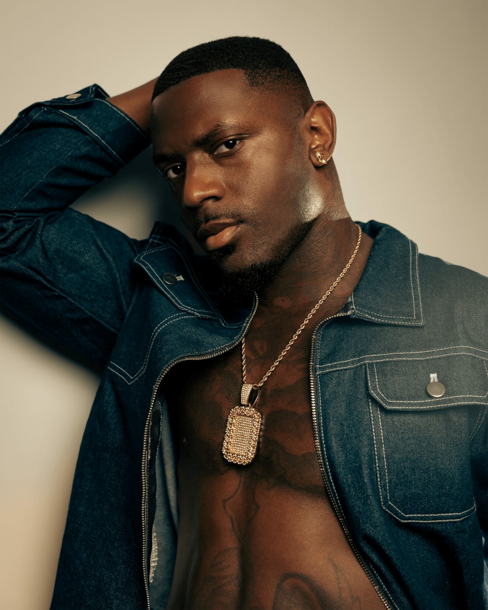 Young man with short hair, wearing a denim jacket and gold chain, posing with hand behind head against plain background.