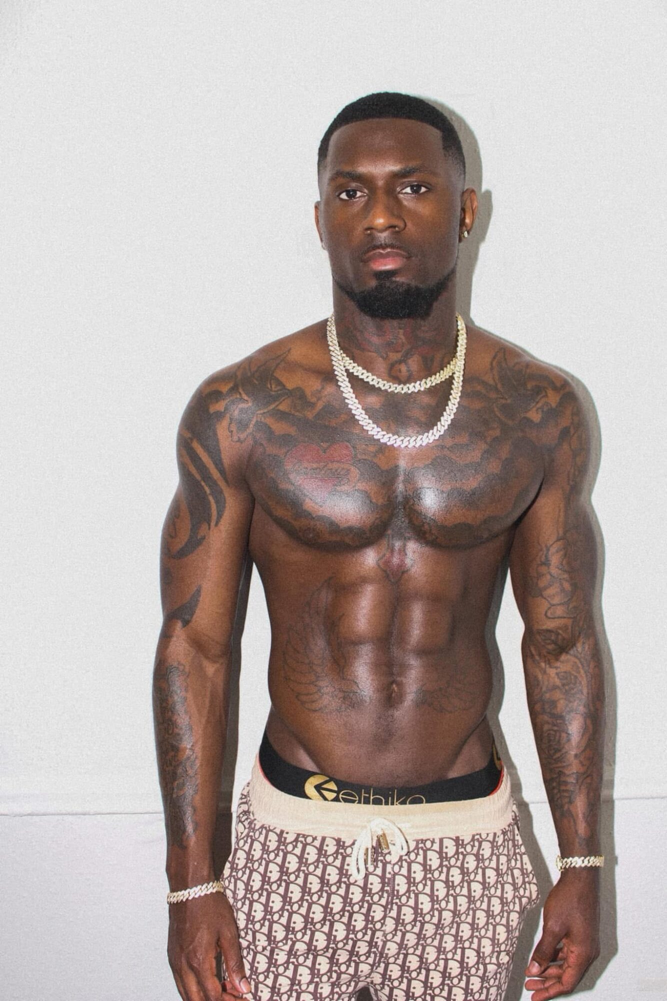Tattooed shirtless man with necklaces and patterned shorts stands against a plain background.