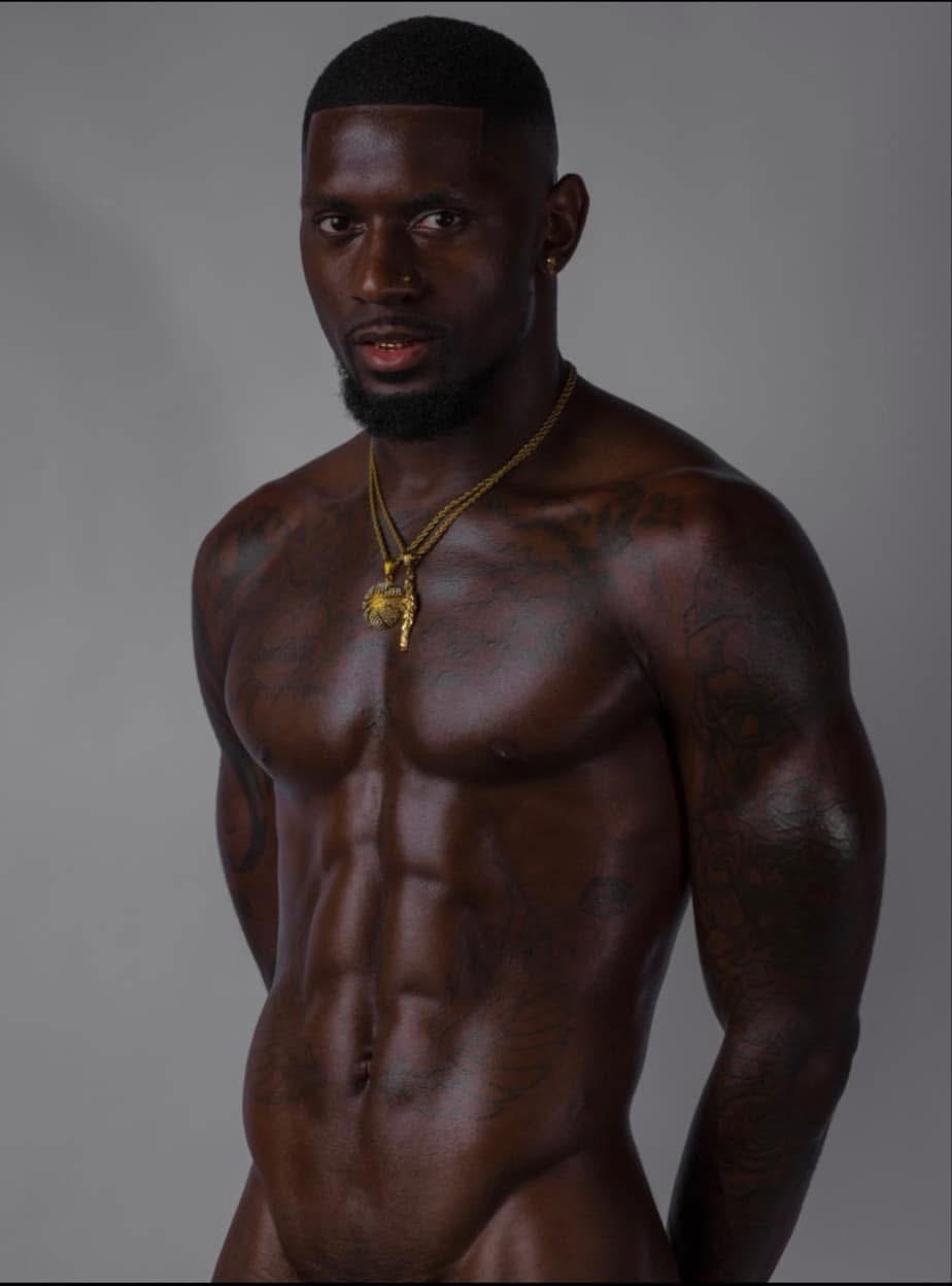 Muscular man with dark skin, short hair, wearing gold necklaces, posing against a plain background.