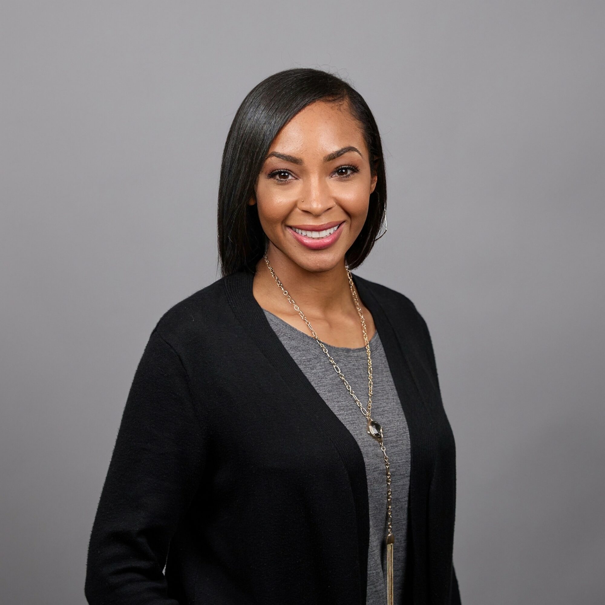 Smiling woman with shoulder-length black hair, wearing a black cardigan and gray top, against gray background.